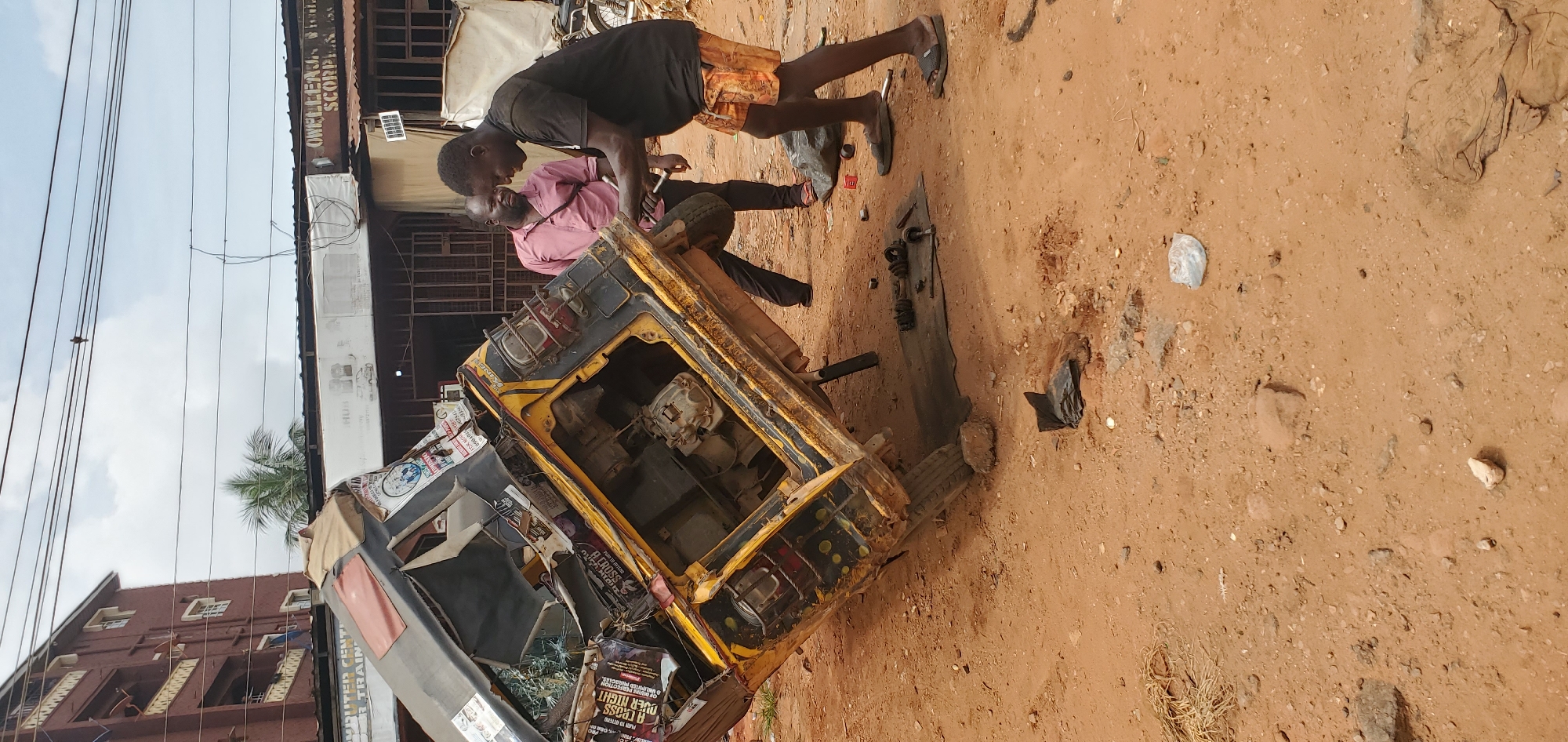 The image captures a scene in Onitsha, Nigeria. Two men are focused on repairing a dilapidated yellow auto-rickshaw (keke) that is missing its front body panels, revealing its engine. One man is wearing a black shirt and orange shorts, while the other is wearing a pink shirt and black pants. They are working on the ground on a tan dirt surface. Several car parts and tools are scattered around them. The background shows buildings. Overhead electric wires are visible against a cloudy sky.