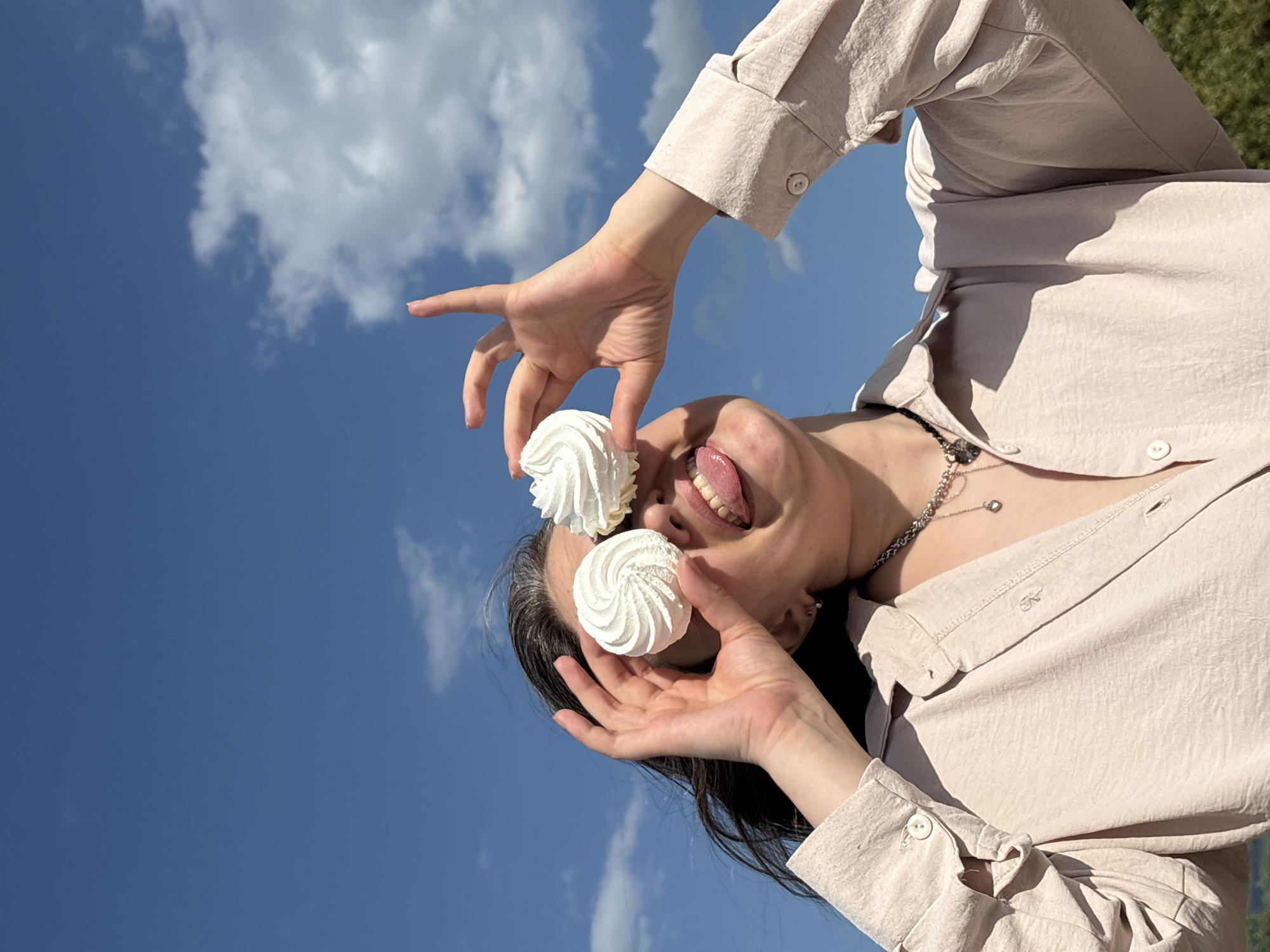 A woman in a tan button-down shirt is turned upside down with the sky and white clouds in the background. She is holding two white meringue cookies, one in front of her eye and one above it, with her tongue sticking out. She is wearing a silver chain necklace. The location is not discernible from the given image.