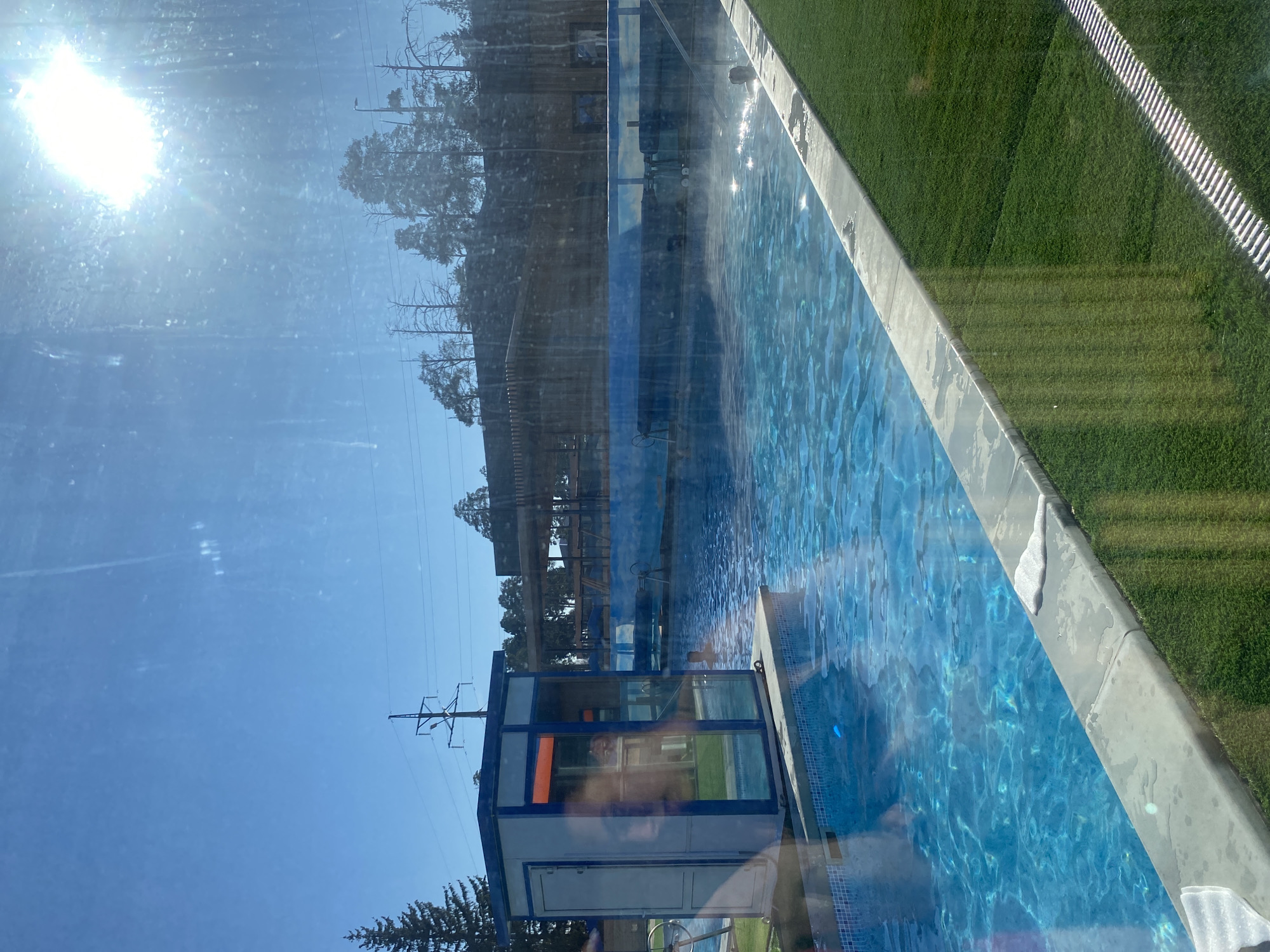 The image depicts an outdoor swimming pool with light blue water. The pool is surrounded by a concrete border on one side, adjacent to green grass and a white-striped surface. Reflections in the water and on a nearby building with a blue and gray exterior are visible. A structure with a glass enclosure is also present. Overhead power lines are silhouetted against a clear blue sky with the sun positioned brightly. Based on the scene's characteristics, the image might depict a location within Votkinsk, Russia, where similar recreational facilities could be found.