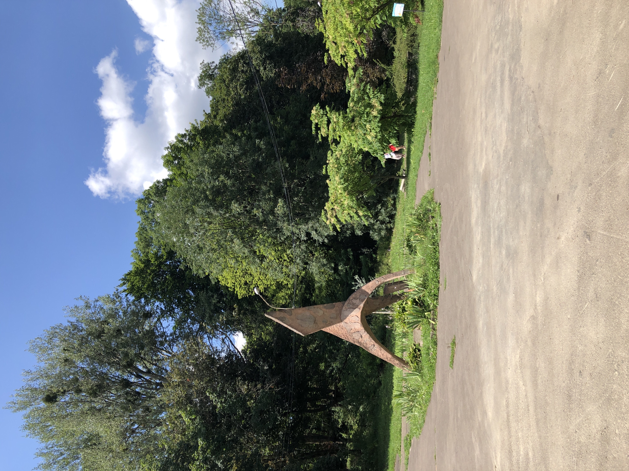 The image depicts an outdoor scene in Lviv, Ukraine, featuring a modern sculpture in a park-like setting. The sculpture, made of reddish-brown metal with a rough texture, has an abstract, angular design with curved elements, resembling a stylized bird or figure in motion. It is positioned on a patch of grass surrounded by a concrete path.
Behind the sculpture, a dense line of trees forms a natural backdrop, with a mix of deciduous trees showing lush green foliage and some darker, possibly coniferous trees. The sky above is bright blue with scattered white cumulus clouds.
In the mid-ground, to the right of the sculpture, there are several smaller, leafy trees with light green foliage. Two individuals are visible walking in this area, one wearing a white top and light-colored pants, and the other in a red top and darker shorts. They appear to be casually strolling through the park.
Further in the background, power lines are visible strung between poles, crossing above the trees. To the far right, a small blue and white informational sign is partially visible among the greenery. The ground in the foreground is a wide expanse of light grey concrete, showing some cracks and weathering. The overall impression is a sunny day in a public park, likely a place for recreation or quiet contemplation.