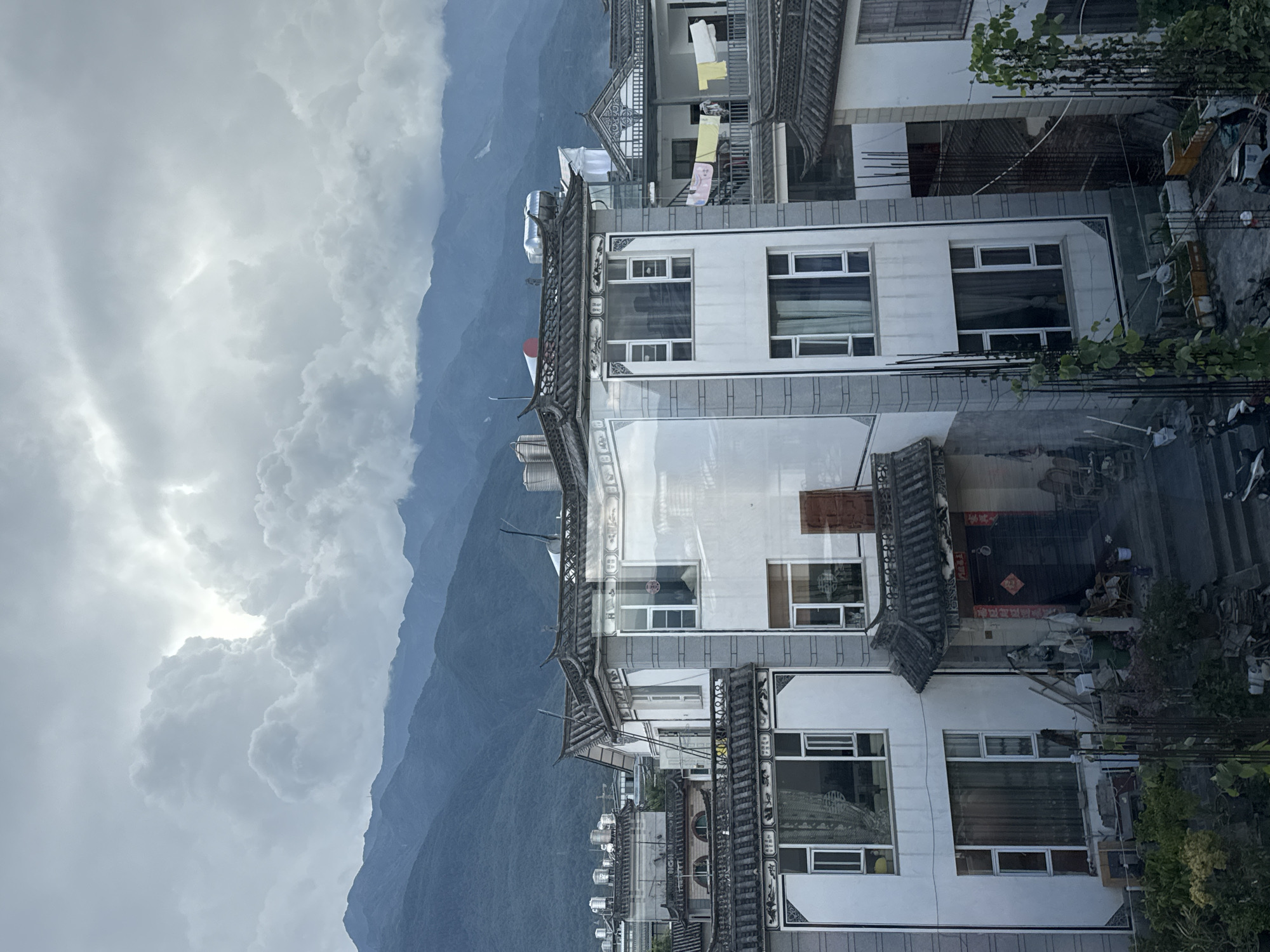 The image depicts a residential area in Dali City, China, characterized by traditional Chinese architecture with dark tiled roofs and white-washed buildings. Mountains loom in the background under a cloudy sky, suggesting a slightly overcast or potentially rainy day. The foreground features multi-story buildings with numerous windows, some of which reflect the sky and surrounding structures. Laundry is visible hanging on a balcony, and various household items, including potted plants, are scattered around the ground level. The overall impression is one of a lived-in, community-oriented urban environment nestled against a natural landscape.
