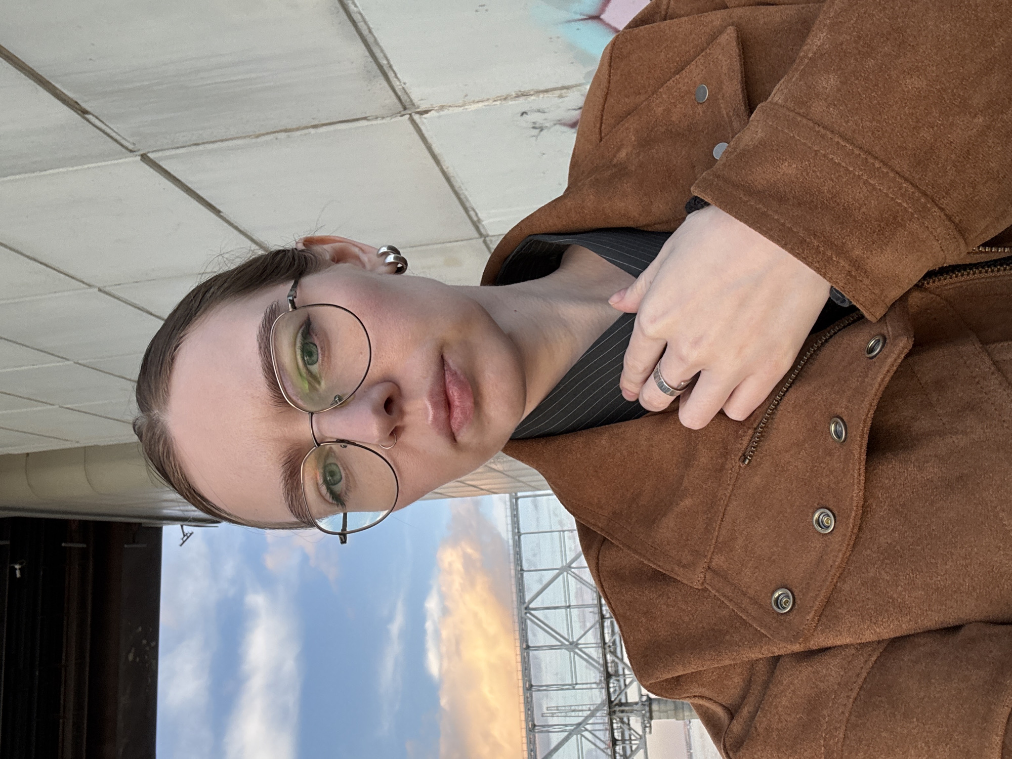 A young woman with light skin and green eyes is centered in the image, wearing round glasses, a dark, striped shirt, and a brown suede jacket. She has a ring on her right hand and a silver earring. The background reveals an urban environment, with a metal structure and a sky transitioning from blue to orange, indicative of either sunrise or sunset. The location is Krasnoyarsk, Russia. A tiled surface is visible behind her.