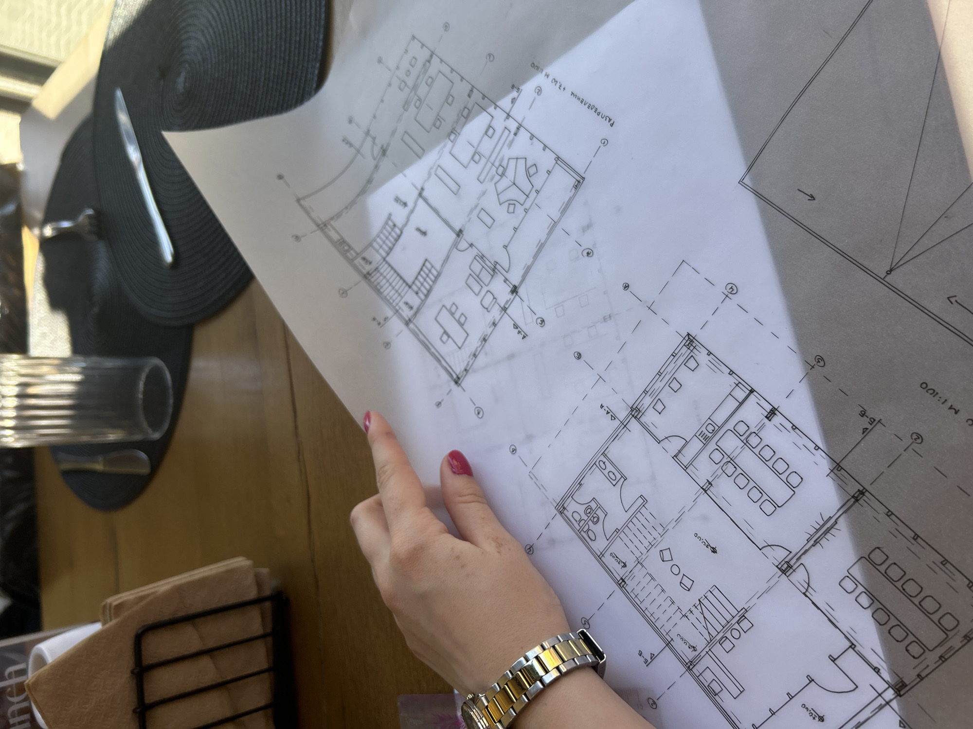 The image shows a person holding a blueprint of a building. The blueprint is white with black lines and text in a foreign language, possibly Bulgarian, as suggested by the text "РАЗПРЕДЕЛЕНИЕ" which is Cyrillic. The blueprint is for two separate floors of a building, with detailed layouts including rooms, stairs, and furnishings. The person holding the blueprint is wearing a gold and silver watch. One hand, with red nail polish, is visible, with the person's wrist below. The background includes a table surface, a black placemat with cutlery, a glass, and a napkin holder with brown napkins. The setting appears to be an outdoor dining area or café, possibly in Sofia, Bulgaria, given the language on the blueprint.