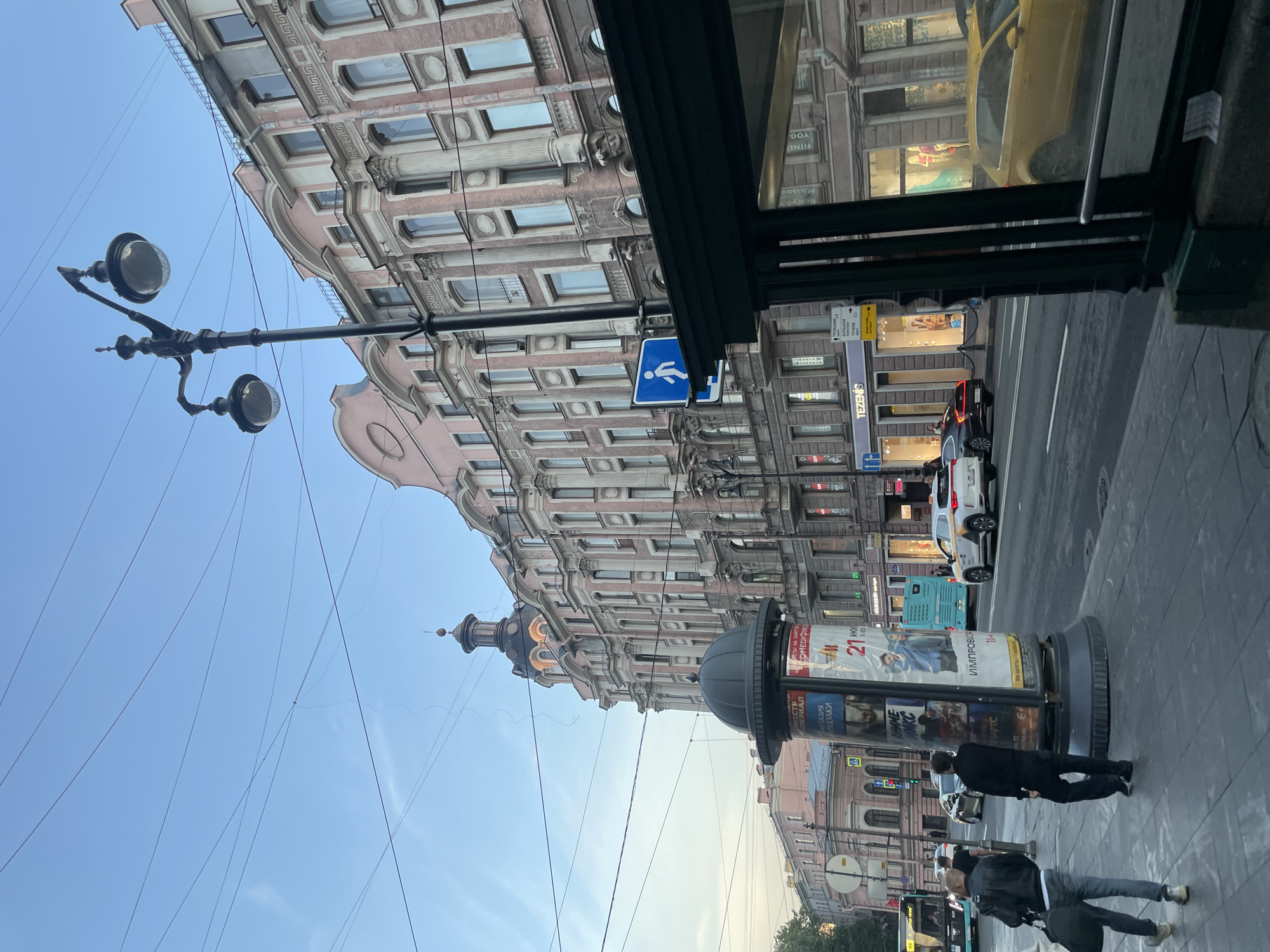 The image depicts a street scene in Saint Petersburg, Russia, likely Nevsky Prospekt, characterized by its grand architecture and bustling urban environment. In the foreground, a pedestrian is walking on the sidewalk, accompanied by another person. To the right, a bus stop shelter with dark green framing and glass panels is partially visible, reflecting the street and buildings. A prominent advertisement column stands on the sidewalk, displaying posters. The street itself features a yellow taxi, a white car, and a turquoise bus, indicating moderate traffic. On the left side of the street, a vintage-style lamppost with two globes stands tall, casting light on the scene. The buildings lining the street are ornate, with pink facades, intricate architectural details, and numerous windows, reflecting the classical style prevalent in Saint Petersburg. Overhead, a complex network of electrical wires crisscrosses the sky, which is a soft blue, suggesting it is either early morning or late afternoon. The overall atmosphere is that of a lively city thoroughfare.