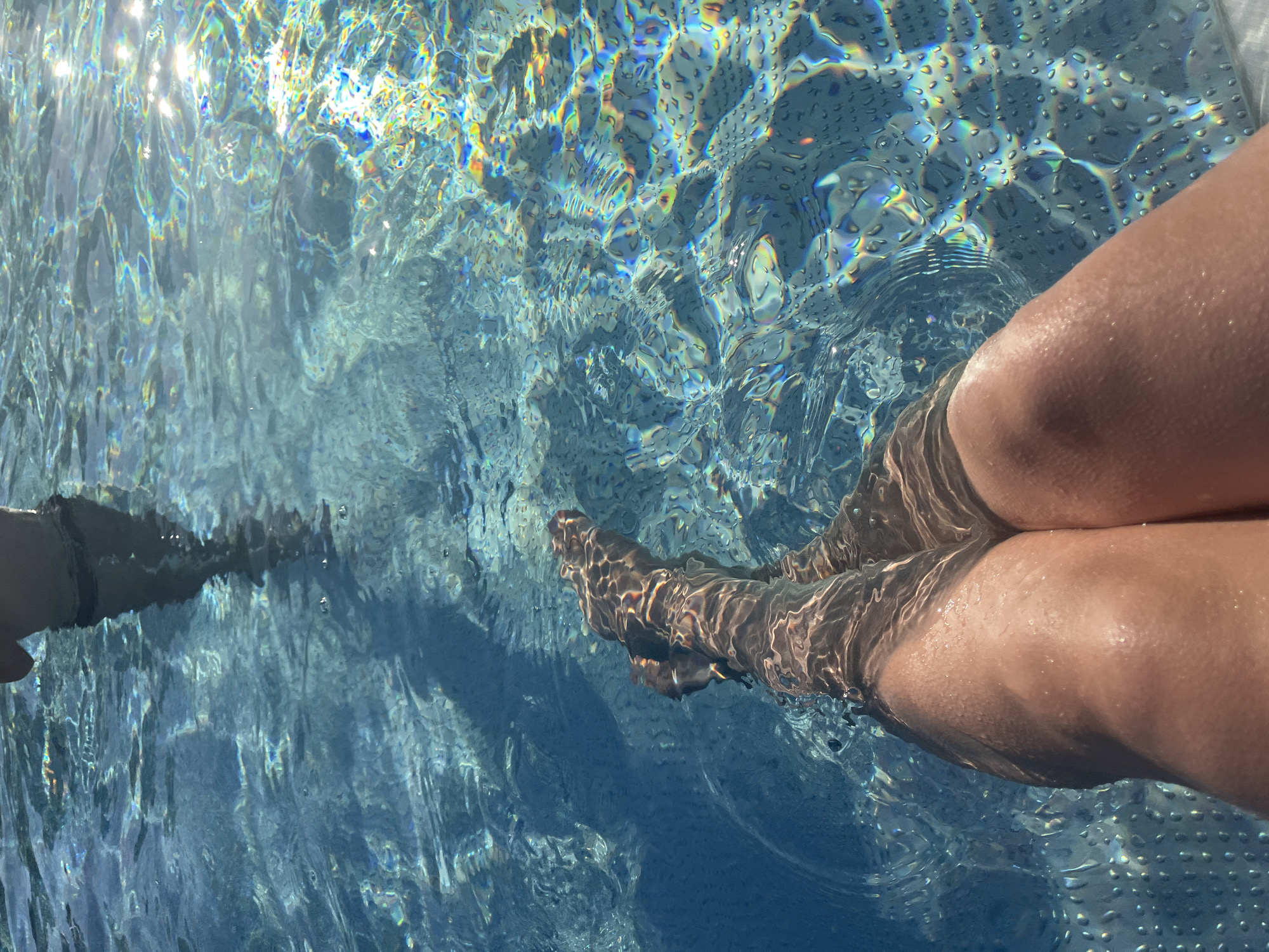 In the city of Unknown, Unknown, a person's legs are submerged in the clear blue water of a swimming pool. The sunlight refracts through the water, creating shimmering patterns of light and color on the pool's surface and the submerged skin. The legs are positioned in a relaxed manner, with one leg slightly bent and crossing over the other. The water is clear, allowing a view of the pool's tiled bottom in some areas. The overall impression is one of a peaceful and sunny day spent enjoying the water.