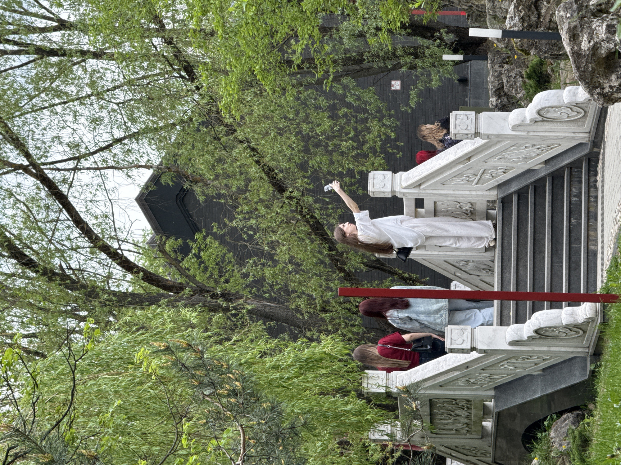 Here is a detailed description of the image:
The image appears to be taken in a park or garden setting in Moscow, Russia, based on the architectural style and foliage. The foreground features a set of ornate, white stone stairs with intricate carvings on the balustrades.
Several people are present on or near the stairs:
- A woman in white clothing stands on the upper part of the stairs, holding up a phone as if taking a photograph.
- Two people are on a lower section of the stairs, and their backs are to the camera.
- One person stands near the top of the stairs.
The stairs are surrounded by lush greenery, including trees with light green foliage. In the background, there is a dark-colored building with a unique roofline, partially obscured by the trees. The overall environment suggests a well-maintained and possibly historical or cultural site. The lighting is soft, indicating either an overcast day or a time of day with indirect sunlight.