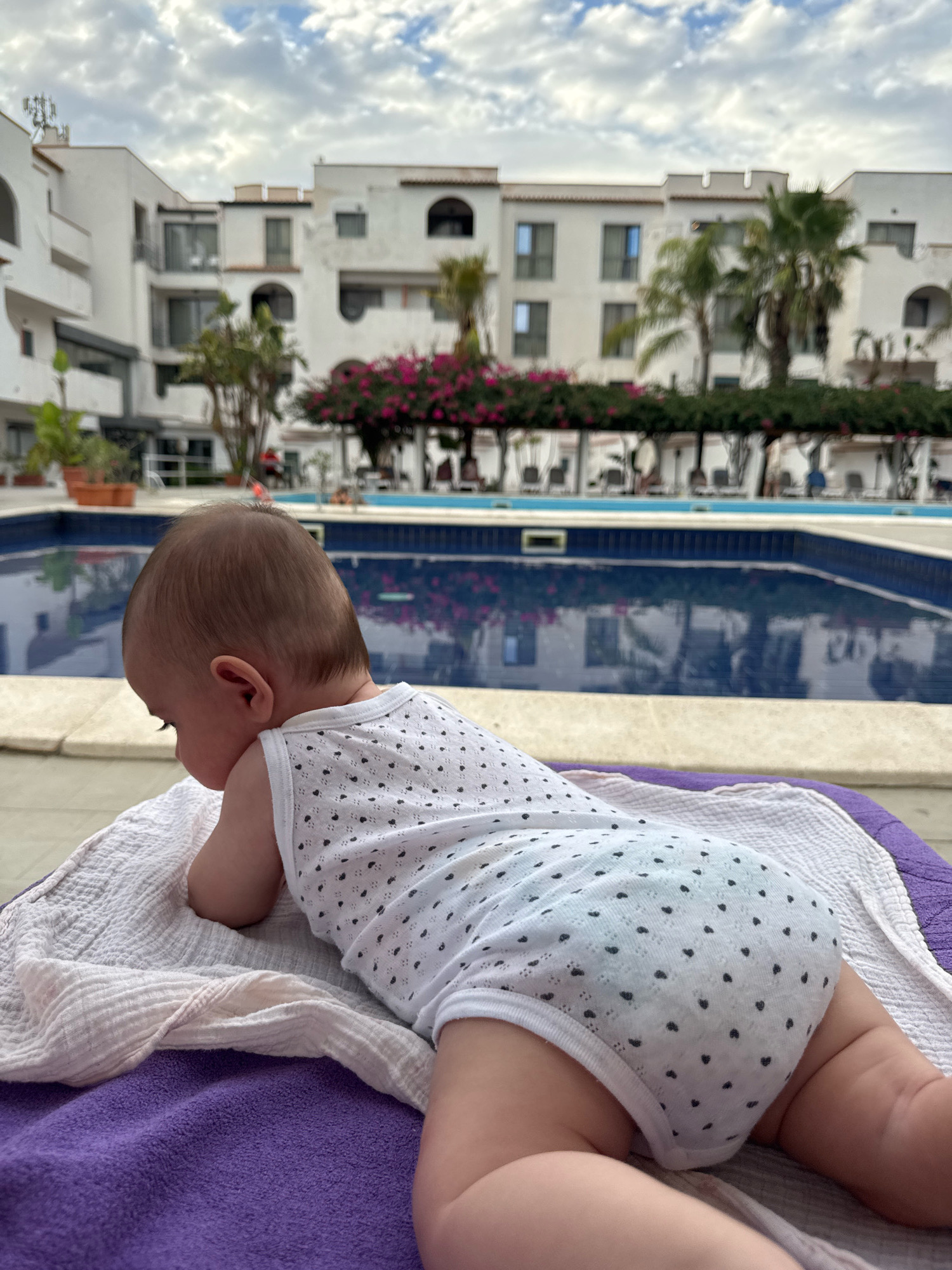 The image shows a baby lying on its stomach on a towel, facing away from the camera. The baby is wearing a white onesie with a black heart pattern. In the background, there is a swimming pool with blue water and reflections of buildings. The buildings are white and appear to be apartments or a resort. There are also palm trees and pink flowers visible. The sky is cloudy. The scene appears to be a vacation or resort setting in Isola delle Femmine, Italy. The lighting suggests it is daytime, possibly late afternoon due to the cloudy sky. The baby's position implies a moment of rest or observation.