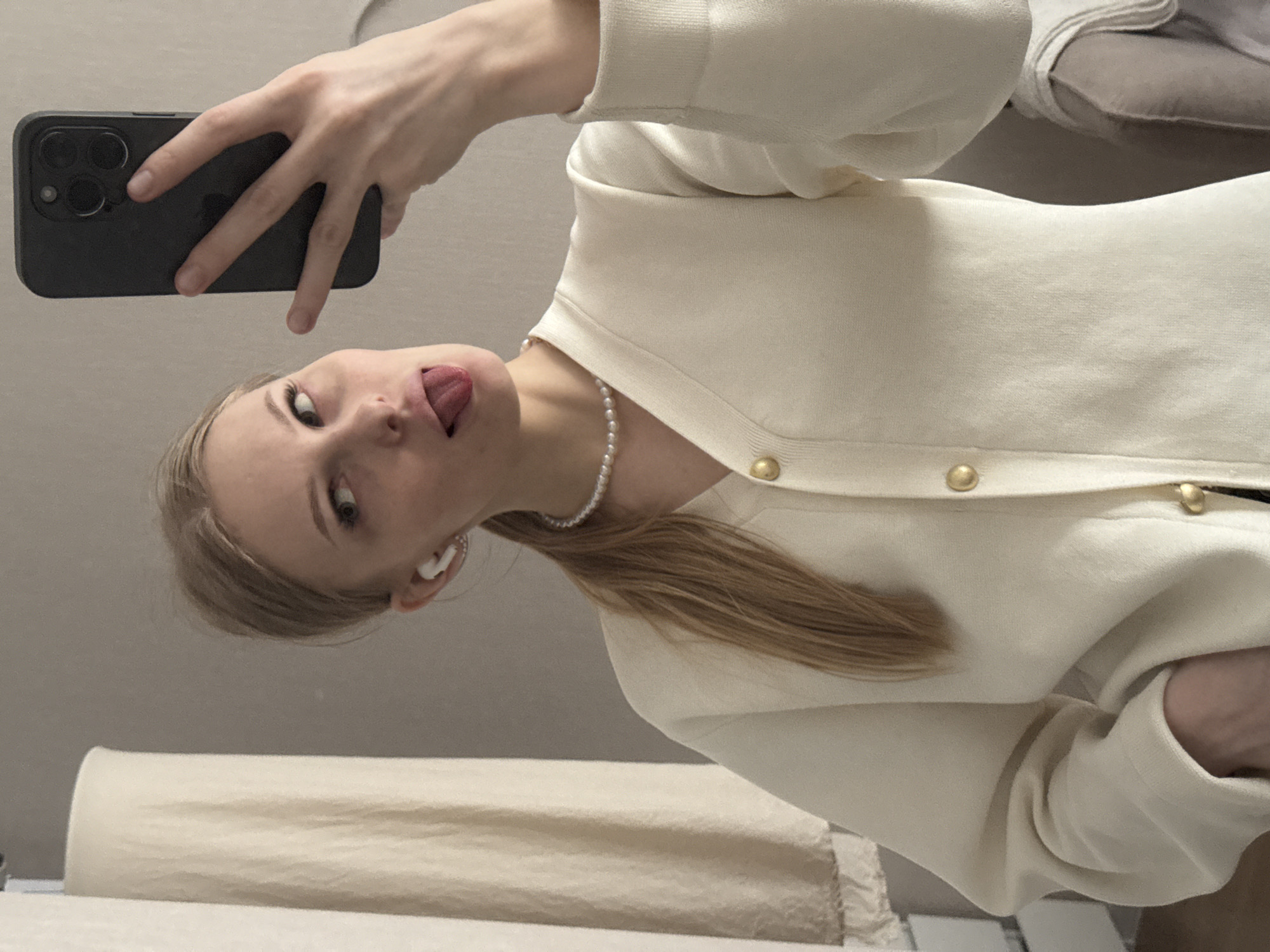 A young woman is holding a black smartphone in her left hand, taking a selfie. She is wearing a pearl necklace and a light-colored cardigan with gold buttons. Her blond hair is pulled back, and she has an earpiece in her ear. She is sticking her tongue out. The background appears to be a room with light-colored walls. The location, Perm, Russia, cannot be determined from the image.