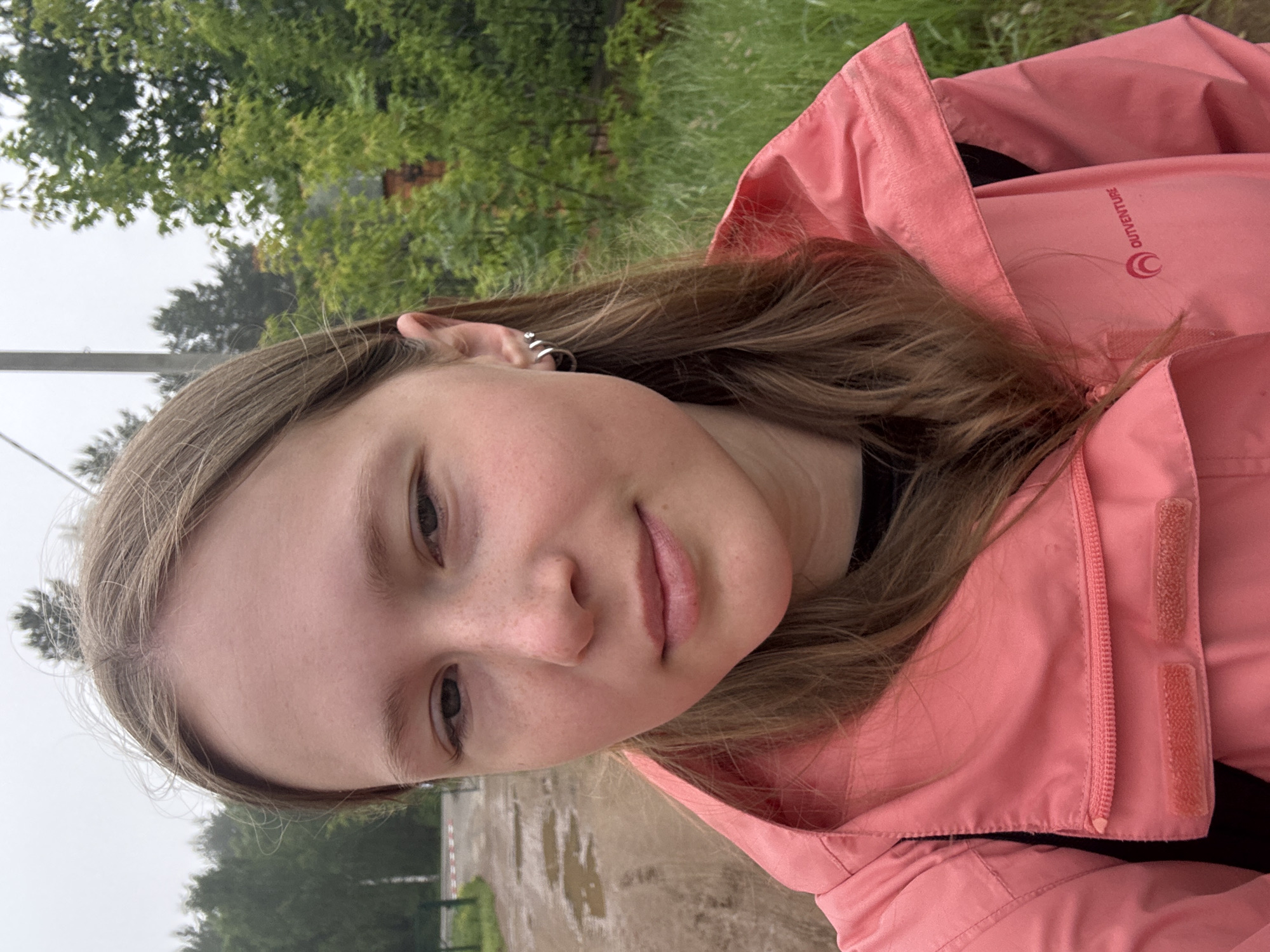A close-up, slightly angled portrait captures a young individual, likely female, outdoors in what appears to be Chaikovsky, Russia. The subject has light brown hair, fair skin, and scattered freckles across her nose and upper cheeks. Her eyes are light-colored, and she wears several small, silver-toned hoop earrings on her visible left ear.
She is dressed in a coral or light pink hooded jacket, which features a zipper and visible velcro strips along the front closure. On the inside flap of the jacket, the brand name "OUTVENTURE" is clearly visible, accompanied by a stylized crescent moon or 'C' shaped logo.
The background reveals a natural setting with lush green trees and foliage filling the upper and side areas. On the ground behind her, numerous puddles are visible on what appears to be a dirt or gravel path or road, suggesting recent rainfall or damp conditions. The sky is overcast, providing soft, diffused lighting.
The subject is looking directly towards the camera with a neutral, calm expression, possibly hinting at a faint, subtle smile. The scene depicts a casual daytime moment under cloudy weather.