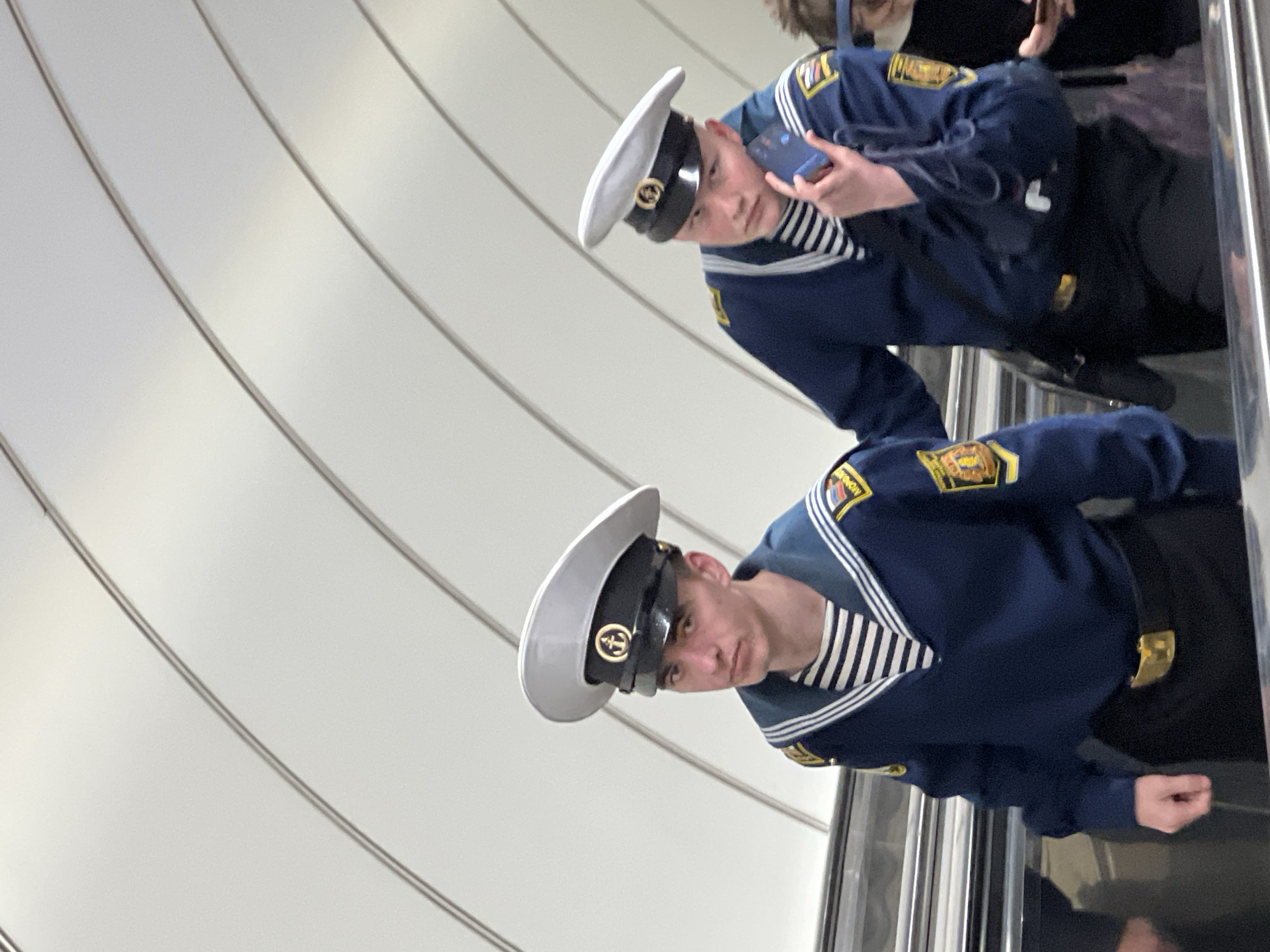 Two young men in Russian naval uniforms are on an escalator. They are wearing white peaked caps with a naval emblem, navy blue sweaters, and striped undershirts. The man at the top of the frame is holding a smartphone. The background features a curved, white-paneled wall, suggesting the location is likely a metro station. Given the presence of naval uniforms, the photo was most likely taken in Saint Petersburg, Russia, a city with a strong naval history.