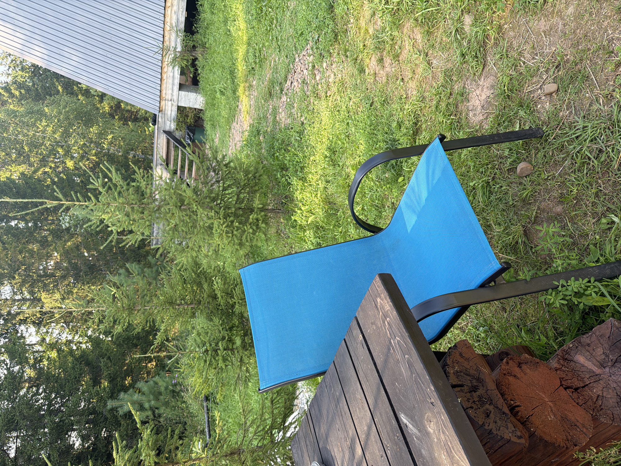 The image depicts an outdoor scene in the city of Kuliki, Russia, featuring a bright blue chair positioned next to a dark wooden table. The table is supported by rough-hewn logs. The ground is covered in lush green grass, with a gravel path visible in the background. A modern, A-frame style building with a corrugated metal roof is situated to the right, accessible by a wooden staircase. Evergreen trees, likely firs, surround the area, creating a natural, wooded ambiance. The lighting suggests it is daytime, with sunlight filtering through the trees, casting shadows on the grass. There are no people visible in the image.