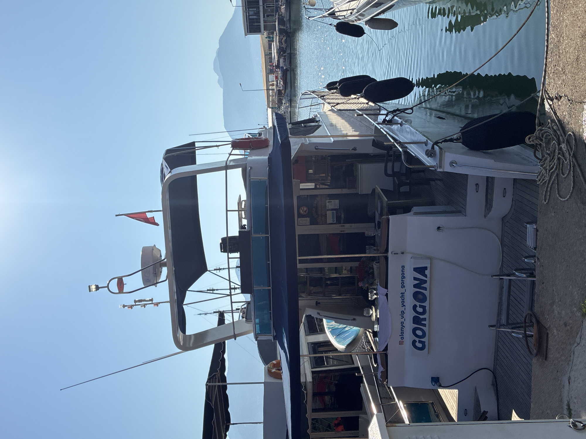 The image shows a white yacht named "GORGONA" docked in the harbor of Alanya, Turkey. The yacht has a blue canopy over the deck and is adorned with several black fenders. On the stern of the yacht, the name "GORGONA" is prominently displayed in blue letters. A table set with glasses and a white tablecloth is visible on the deck. The background features other boats, a waterfront promenade, and a mountain range under a clear blue sky. The sun is shining brightly, casting reflections on the water. There are no people visible in the image.