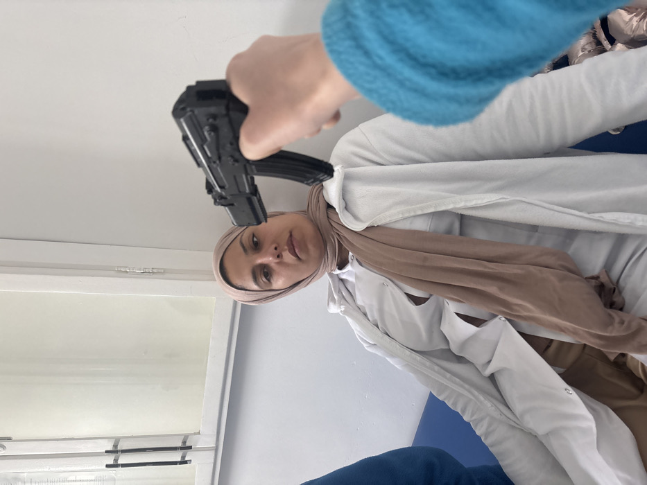 A woman wearing a hijab and a white coat is pictured from a low angle. A black toy gun is pointed at her head. An arm, clad in a blue sweater, holds the gun. The woman looks directly at the camera with a neutral expression. The background includes a window and a blue cushioned area. The image has been rotated to an odd angle.