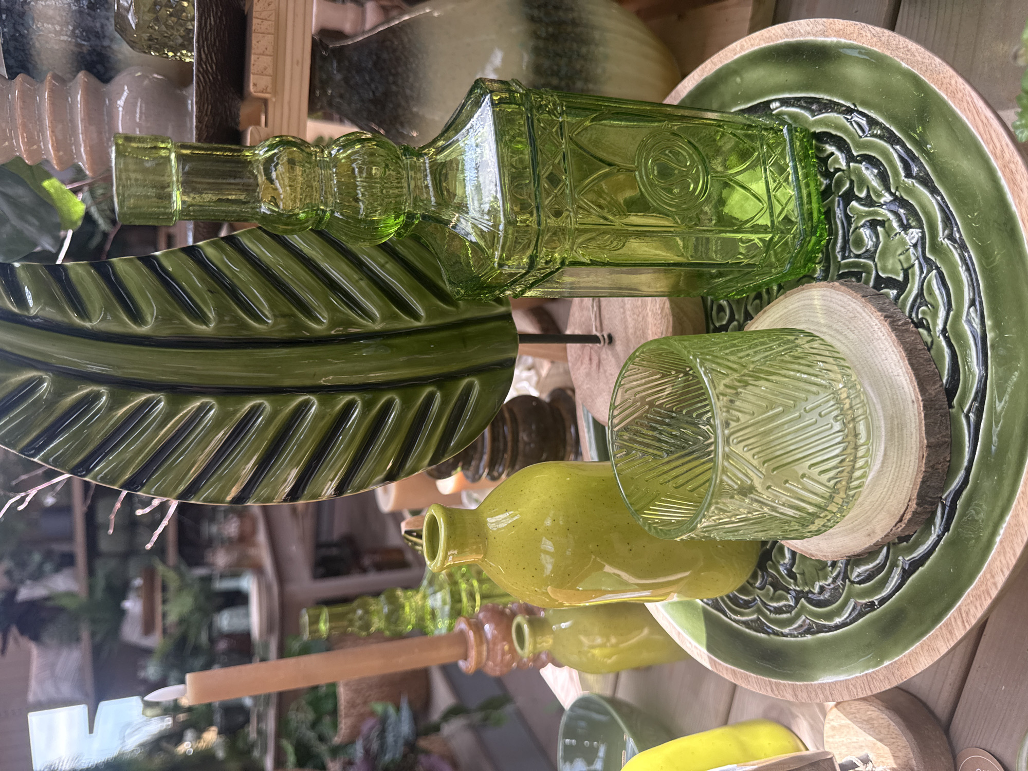 A tabletop display of green glass objects is presented. In the foreground, a green, intricately carved plate rests on a wooden tray. On the plate are two green glass items: a cylindrical glass with a textured pattern, supported by a slice of wood, and a tall, square-shaped bottle with decorative relief. To the left of these items, a smaller, bulbous green bottle is visible. Behind these, a tall, slender green glass bottle stands. Further back, a large, green, leaf-shaped decorative object is prominent. A cream-colored candle on a wooden base is to the left of the leaf. The background is blurred, showing shelves with various decorative items, including ceramic vases and plants, suggesting an indoor retail or decorative setting. The scene is indoors, possibly in Fritzlar, Germany, given the prompt. No people are present in the image.