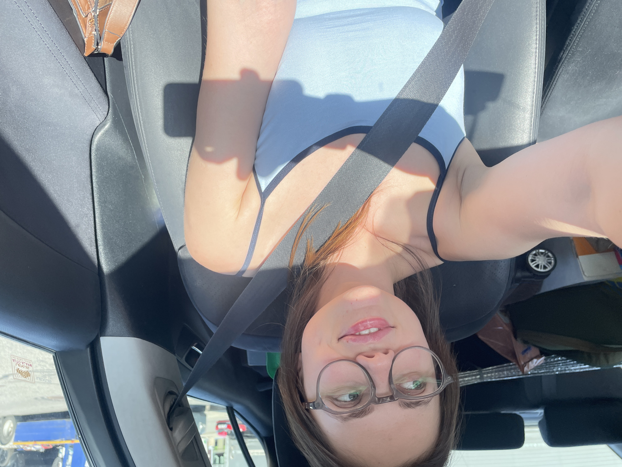 The image shows a woman in a light blue tank top and glasses wearing a seatbelt in a car. The interior of the car is visible, including the dashboard and seats. The woman is looking at the camera. Through the window, there is a trailer and a gas station. It is not possible to ascertain that the image was taken in Jihlava, Czechia based on the visible information.