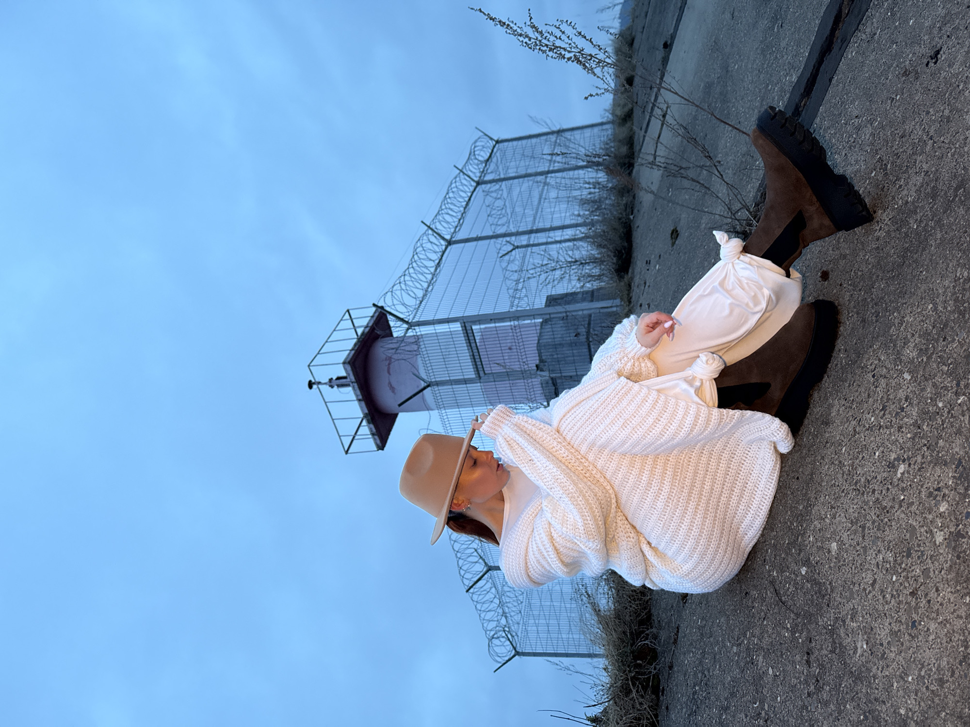 An outdoor, full-body portrait features a woman seated on a rugged concrete surface, with her legs extended forward. She is styled in a cozy, fashionable ensemble, wearing an oversized, chunky knit cream or white sweater, light-colored pants, and dark brown combat-style boots with thick black soles. A light tan wide-brimmed fedora-style hat covers most of her head, with a glimpse of dark, possibly reddish-brown hair peeking out. She is captured in a contemplative pose, looking downwards and to her left, with one hand gently resting on the brim of her hat. Her fingernails are painted a light nude or pink.
The setting is an industrial or semi-abandoned outdoor area, likely in Chaikovsky, Russia. In the background, a prominent chain-link fence topped with coils of razor wire extends across the frame. Behind the fence, a tall, reddish-brown industrial tower or structure with a small platform and machinery at its peak is visible. The ground consists of textured concrete, with sparse, dry vegetation growing nearby. The sky is a clear, light blue, suggesting soft, even lighting, indicative of either early morning, late afternoon, or an overcast day. The overall impression is a blend of modern fashion against a stark, utilitarian backdrop. No text is visible in the image.