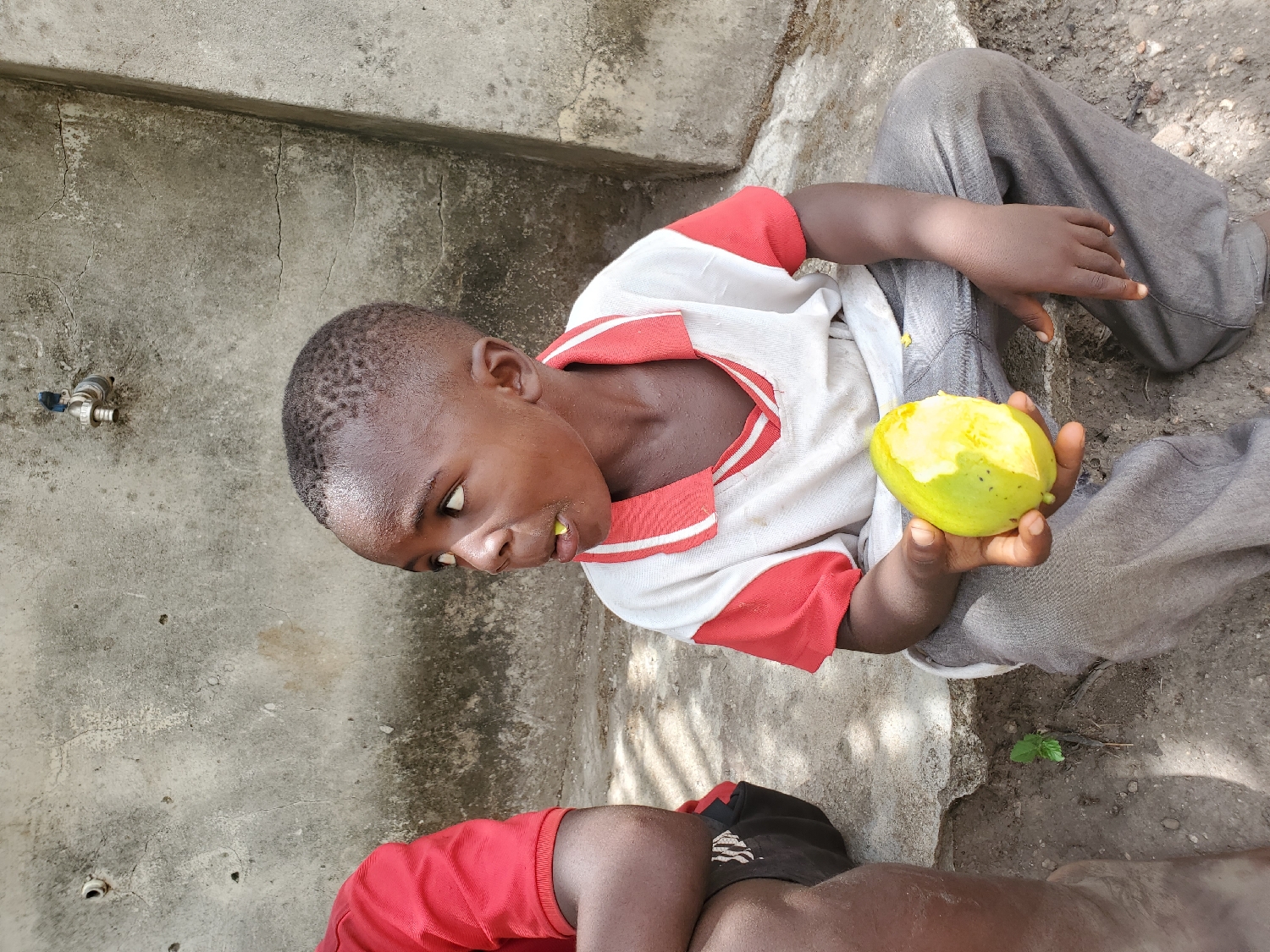 A young boy, appearing to be of primary school age, is the central subject of this outdoor, eye-level shot, taken in Mararaba, Nigeria. He is seated on the ground, leaning back against a concrete surface. He wears a white polo shirt with red stripes on the collar and shoulders, and grey trousers. In his right hand, he holds a partially eaten, light green and yellow mango, some of its flesh visible around his mouth, indicating he is actively consuming it. His expression is neutral as he looks slightly upwards and to his right, towards the camera.
In the upper left background, a small, silver-colored water tap or faucet is mounted on a textured, light grey concrete wall. The ground beneath him transitions from concrete to packed dirt on the right. The lower left foreground shows the arm and leg of another person, likely another child, dressed in a red shirt and black shorts, partially obscured by the main subject.
The bright, direct lighting suggests it is daytime, likely sunny. The concrete surfaces show some cracks and wear, while the dirt ground appears dry. There is no visible text in the image. The scene depicts a simple, everyday moment of a child enjoying a mango in an outdoor, informal setting.