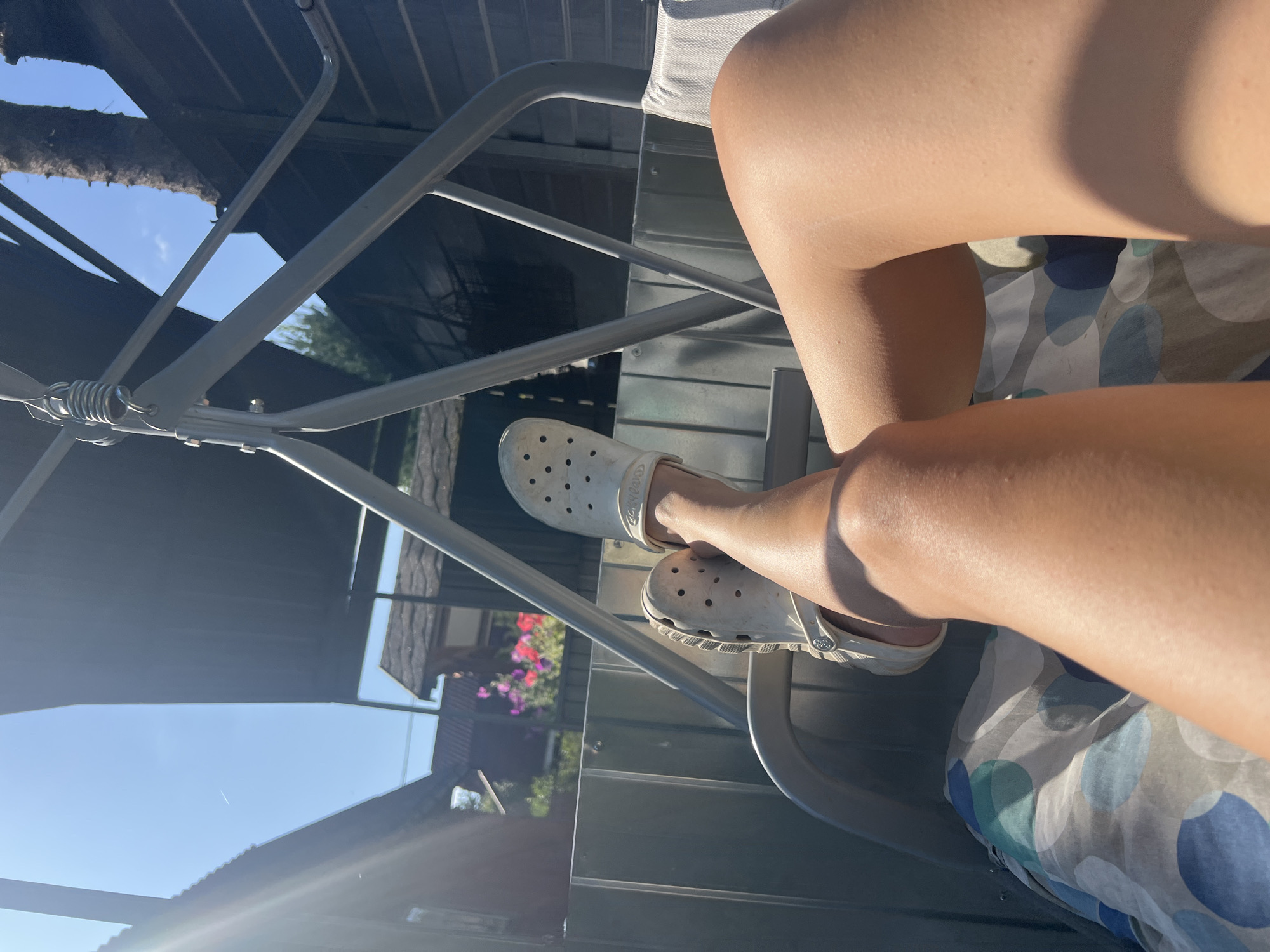The image shows a close-up view of a person's legs, clad in white Crocs, resting on a striped cushion on a swing. The swing is gray metal with a black fabric canopy. In the background, there are houses with dark roofs, some trees, and vibrant pink and red flowers. The scene appears to be set in a backyard on a sunny day, possibly in the dacha settlement of Роса Снт, Russia, given the context provided. The sunlight casts shadows across the legs and the swing.