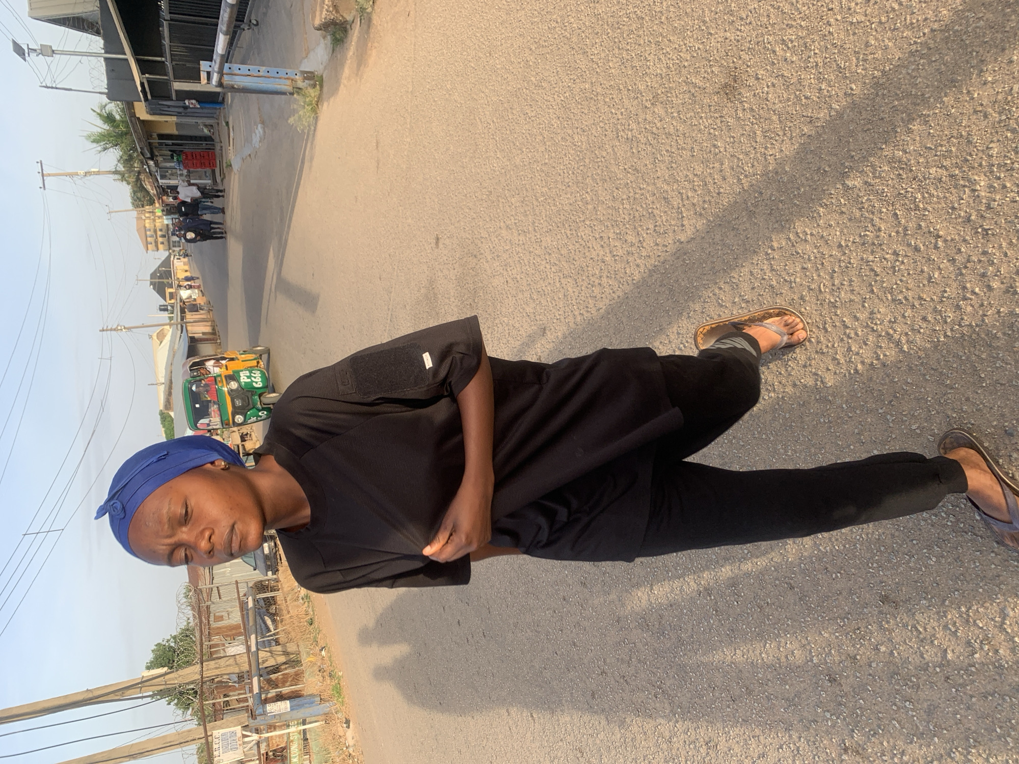 The image shows a woman standing on a paved road in what appears to be a residential or commercial area in Jos, Nigeria. She is wearing a dark-colored outfit, a blue head covering, and sandals. Her shadow stretches out on the road beside her. The background reveals structures, utility poles, and what appears to be a motorized tricycle vehicle (keke) commonly used for transport in Nigeria. Other people are faintly visible in the distance. The overall scene suggests an outdoor setting in daylight.