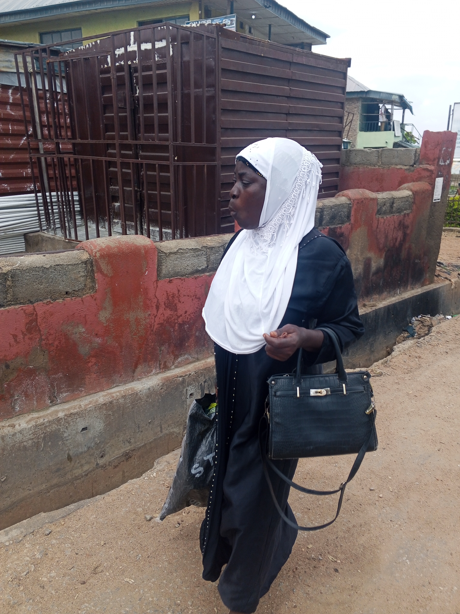 A medium shot captures a Nigerian woman of apparent African descent, wearing a white hijab and a long black abaya, walking to the left in Zango-ndaji, Nigeria. She holds a black alligator-skin handbag with gold hardware in her right hand and a black plastic bag in her left. The woman is positioned in front of a wall with a weathered red facade. Behind the wall, a large, dark brown metal structure, possibly a shipping container or storage unit, is visible. The ground is composed of dirt and gravel. The background shows a building with a yellowish exterior and a roof extending outwards. The sky is overcast.