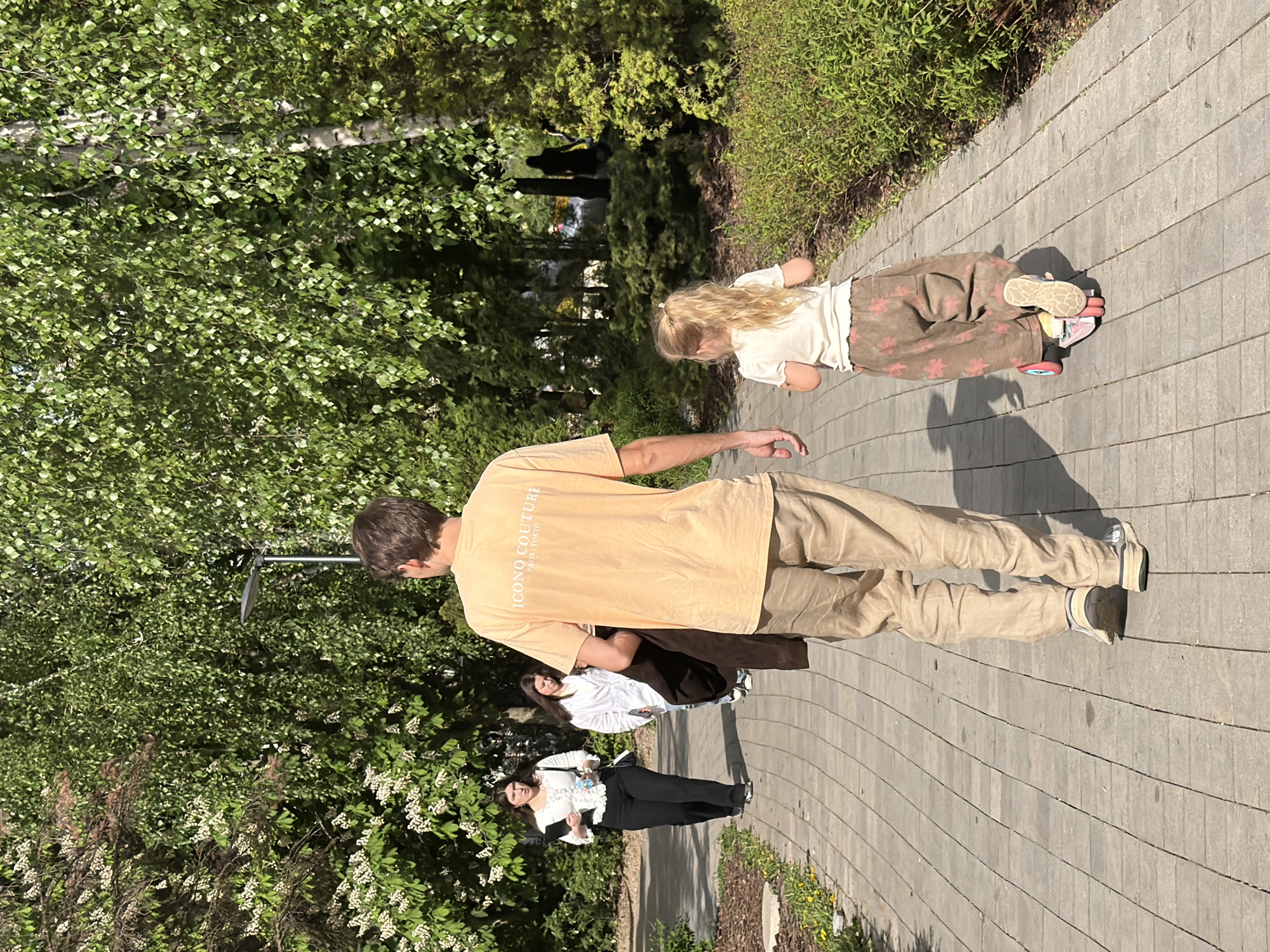 A daylight outdoor scene shows a paved walkway in a park setting. A man in a peach-colored t-shirt with the words "ICONO COUTURE PARIS TOKYO" and beige pants walks along the path. Behind him are two women, one wearing a white shirt and jeans and the other wearing a black and white shirt and black pants. In front of the man, a young girl lies on a pink skateboard. The background includes trees and foliage. Based on the architecture and general appearance of the park, it appears to be located in Moscow, Russia.