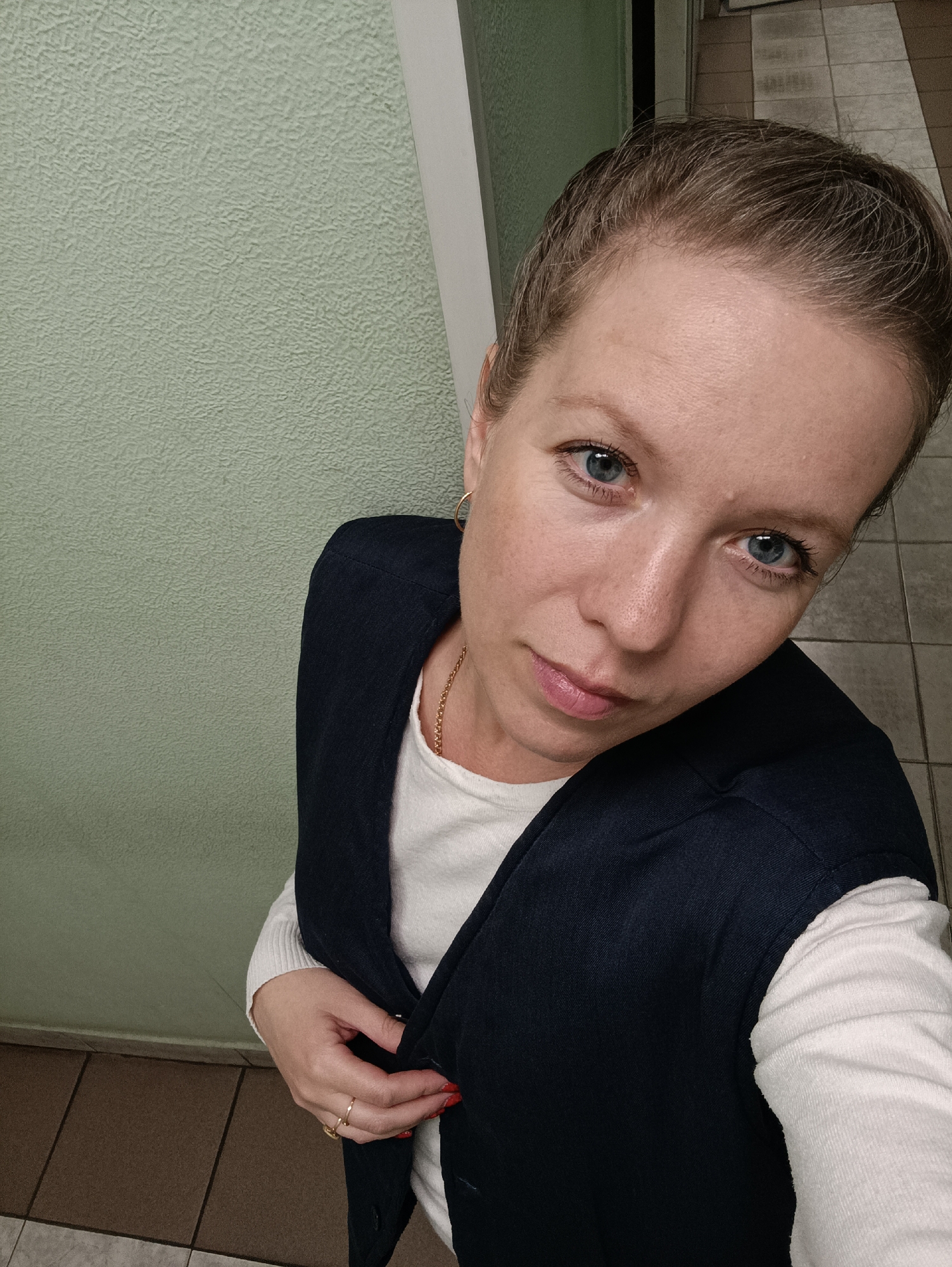 A woman with fair skin, short blonde hair, and blue eyes is taking a selfie in a room. She is wearing a white long-sleeved shirt under a navy blue vest. She has gold hoop earrings and a gold chain necklace. Her nails are painted red. The room has a light green wall with a textured surface. The floor is tiled in shades of brown and beige. The location is Chaikovsky, Russia.
