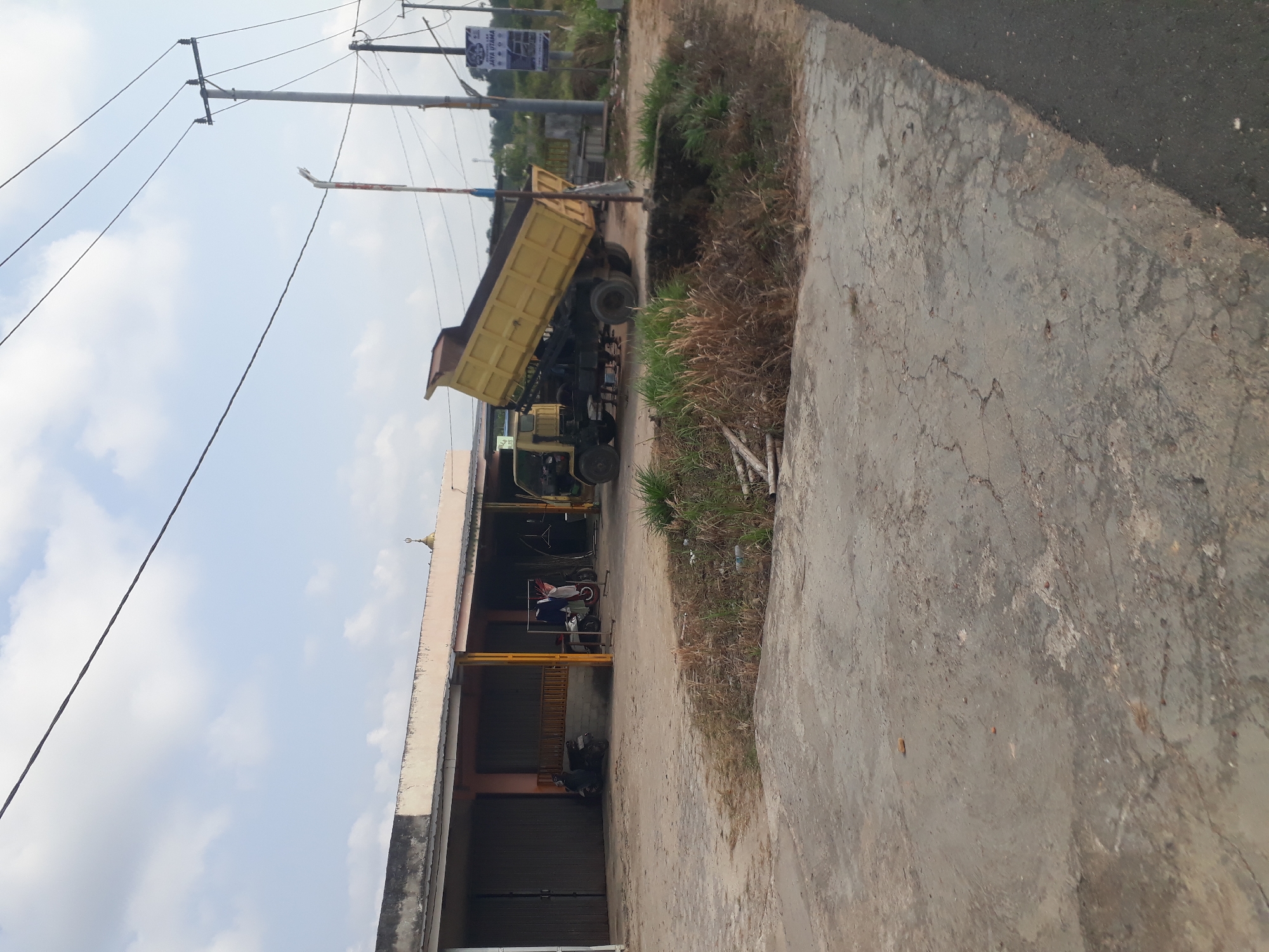 In Desa Balun Ijuk, Indonesia, a yellow dump truck with its bed raised is positioned on a dirt and concrete surface. In the background, a long, single-story building with a light-colored facade and dark gray roll-up doors lines the area. To the left of the building, a dark-colored scooter is parked, and to the right, a white scooter is also visible. A rack with clothes hangs on the right side of the building. Power lines stretch across the sky above the scene, supported by utility poles. A sign with Indonesian text and images is mounted on one of the poles. Dry grass and some scattered debris are present on the ground in front of the building and truck. The sky is partly cloudy.
