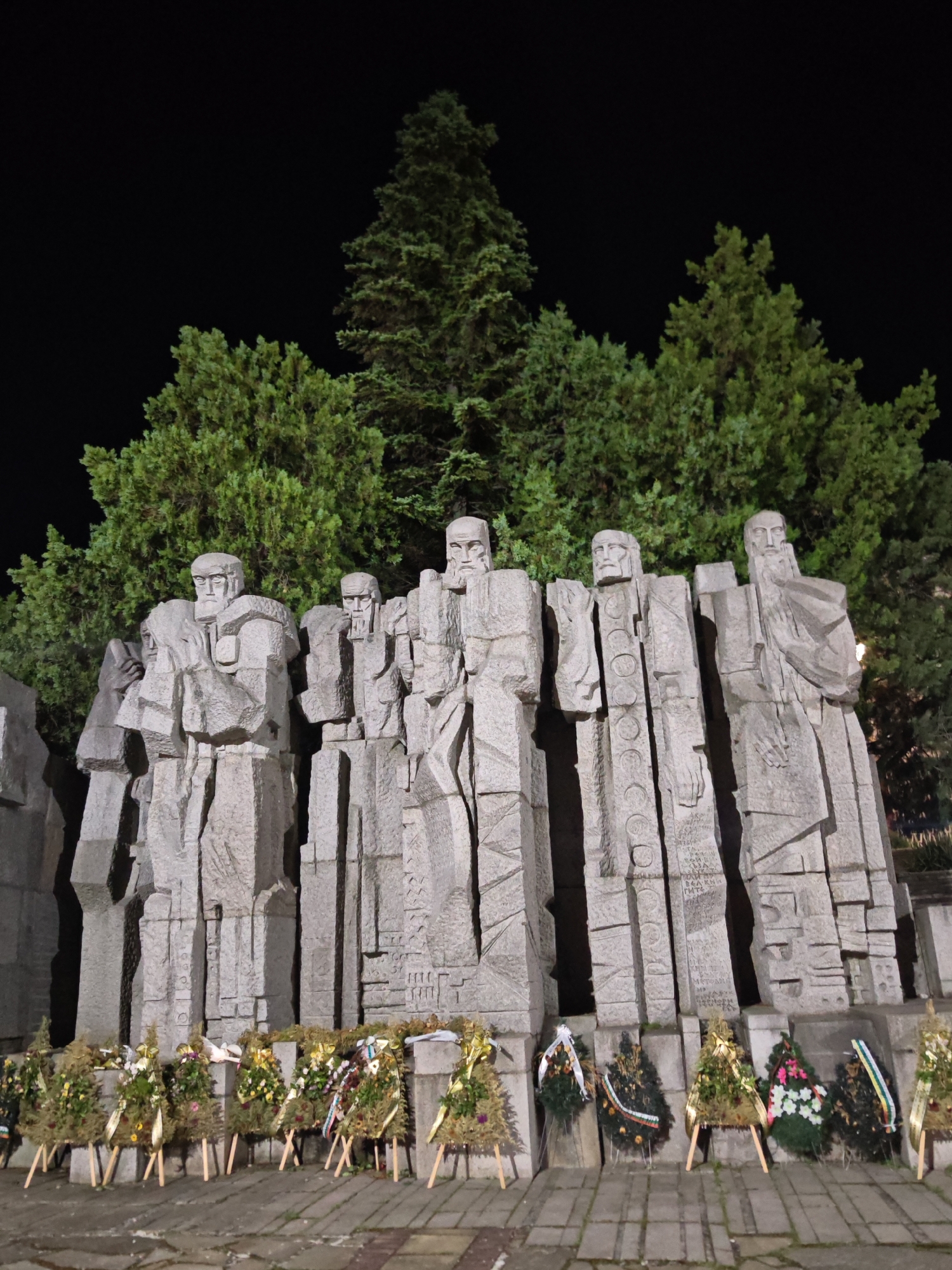 This nighttime image depicts a large, monumental stone sculpture situated in Veliki Preslav, Bulgaria. The central subject is a series of at least seven stylized, blocky male figures carved from light-colored stone, standing in a continuous row. Each figure features a prominent beard and abstract, geometric facial and body features, imparting a solemn, heroic quality. The figures are integrated into a larger, textured stone wall structure.
Behind the monument, a dense formation of evergreen trees, possibly conifers, is clearly visible and illuminated, creating a vibrant green backdrop against the stark blackness of the night sky.
In the immediate foreground, along the base of the stone monument, a collection of approximately ten memorial wreaths and floral arrangements is displayed. Many of these wreaths are supported by small, light-colored wooden easels. The wreaths primarily consist of green foliage, interspersed with various colorful flowers and ribbons, some of which exhibit patterns and colors suggestive of national affiliations, such as the Bulgarian flag (white, green, red).
The ground in the foreground is a flat, paved surface composed of rectangular stone or concrete slabs. The entire scene is lit by artificial light sources, which illuminate the monument, the wreaths, and the trees, while the upper sky remains in complete darkness.