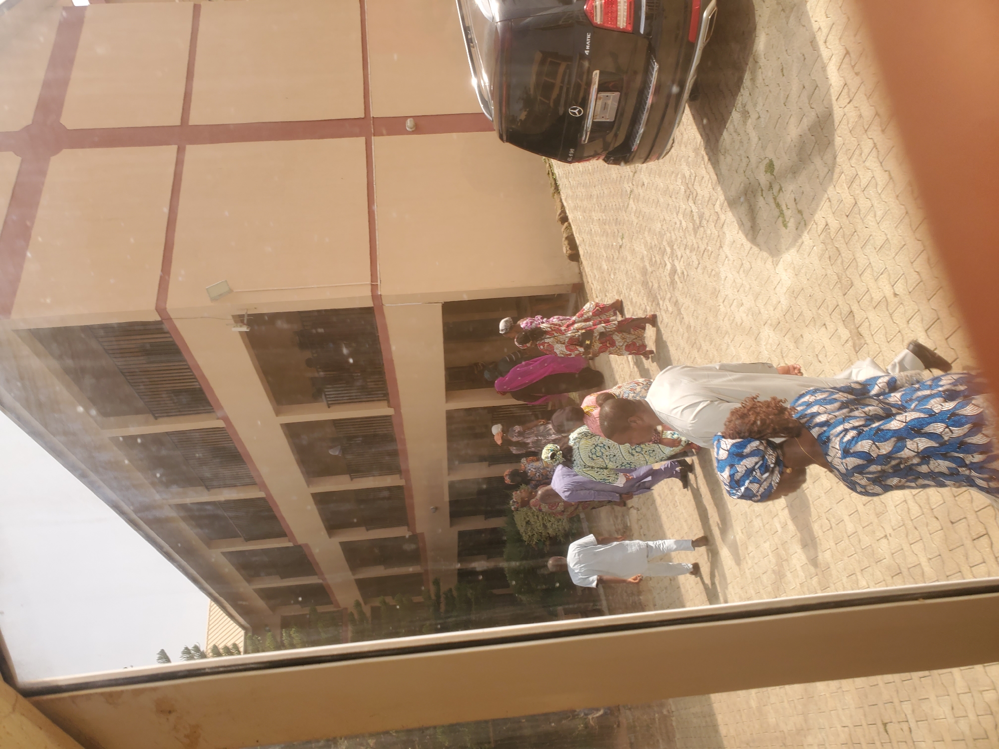 The image depicts a group of people gathered on a paved walkway in front of a two-story building, likely located in Jos, Nigeria. The building's facade is a light tan color with darker tan trim along the edges of each floor and vertical accents. Each floor has a series of balconies with dark railings. A black Mercedes-Benz GL-Class SUV is parked on the right side of the paved area. Several individuals are visible in the foreground and mid-ground, most of whom are wearing traditional African attire in various colorful patterns. One person in the foreground on the right is wearing a blue and white patterned headscarf and dress. Another individual in the mid-ground on the left is wearing a white traditional outfit. The ground is paved with interlocking beige brick pavers. The scene appears to be outdoors, under daylight.