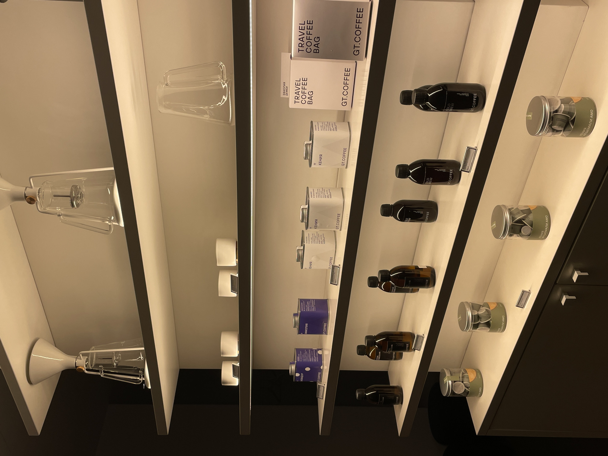 The image shows a shelf display of coffee products, likely in a retail setting. The shelves are white and angled, containing various items including boxes labeled "TRAVEL COFFEE BAG" and "GT.COFFEE", cans with names like "КЕНИЯ" (Kenya) and "АЛШЕМАЛИ АМО", and bottles labeled "COLD BREW". There are also glass coffee brewing apparatuses on the leftmost shelves. The overall impression is a curated display of coffee-related merchandise. Based on the Cyrillic text on the products, the location is likely in Moscow, Russia.