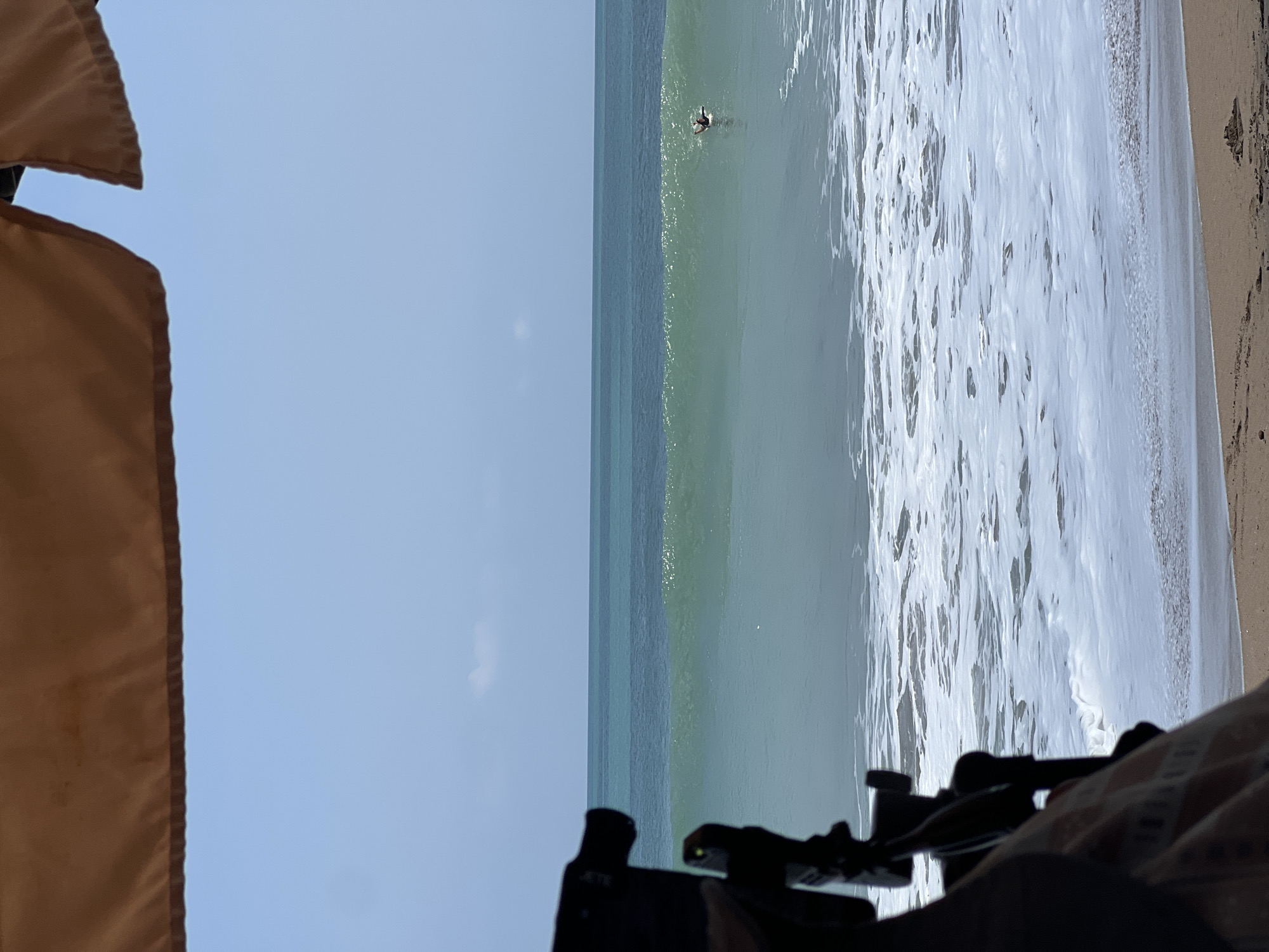 In Kuta, Indonesia, a serene beach scene unfolds under a pale blue sky. In the foreground, the bottom half of a dark object, likely a camera or tripod, is partially visible, casting a shadow on a patterned cloth. The ocean stretches out, with a clear horizon dividing the water and sky. A gentle wave is forming, its translucent green surface catching the light. In the distance, a lone surfer is positioned on a surfboard, seemingly waiting for or riding the wave. The water transitions from a lighter turquoise near the shore to a deeper blue further out. The beach itself is composed of fine, light brown sand, with some small debris scattered on its surface. The overall atmosphere is peaceful and tranquil.