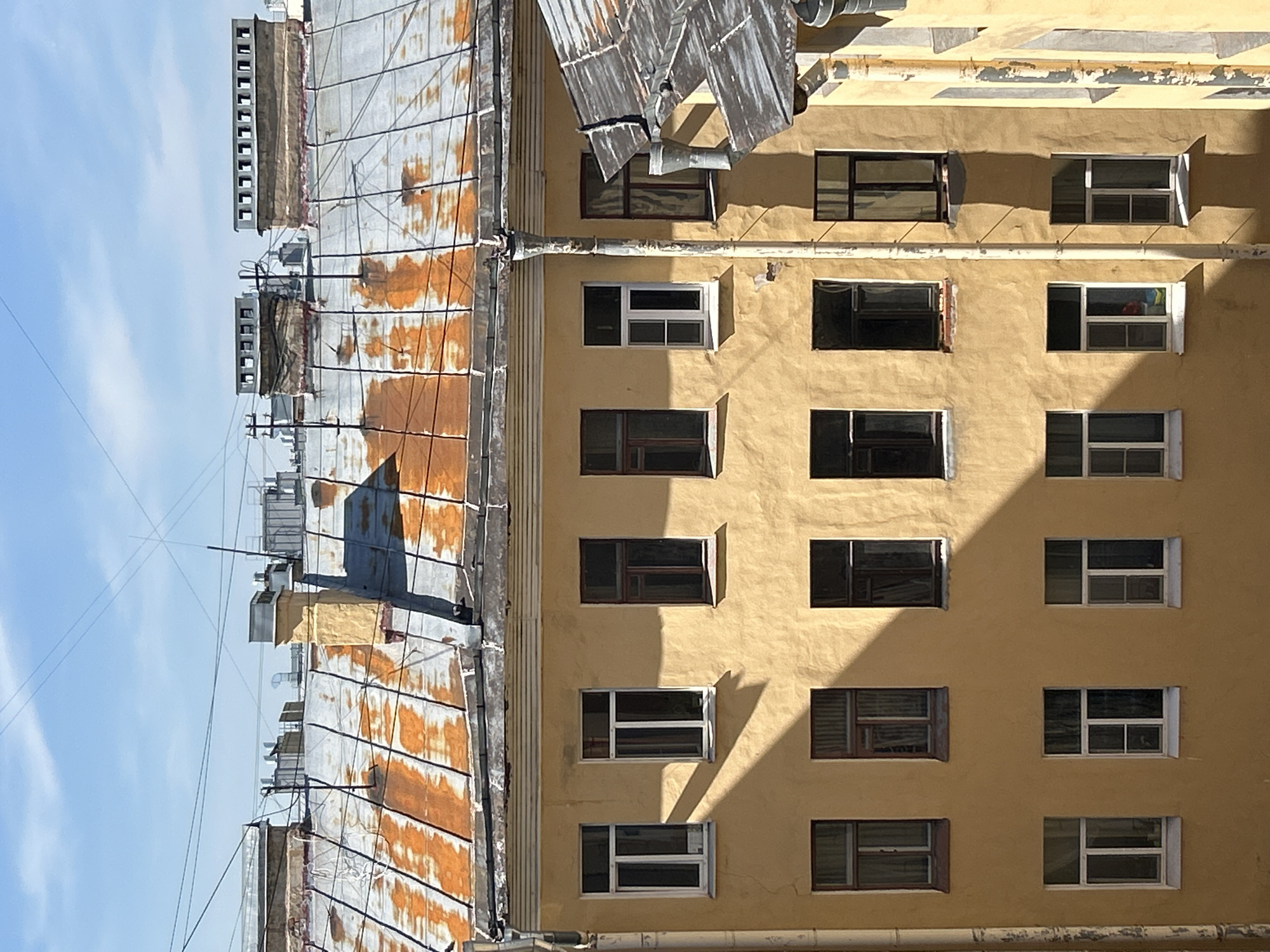 An eye-level outdoor shot depicts the architecture of a building in Saint Petersburg, Russia, during daylight under a partly cloudy sky. The building is viewed from below, looking upward.
The primary subject is a multi-story building with a yellow-painted exterior and a rusted, corrugated metal roof. The facade features multiple windows in a grid-like pattern, some open and some closed, displaying dark interiors. The building appears to be in a state of disrepair, with paint peeling in areas.
The roof shows significant signs of rust and wear, with some sections of the corrugated metal appearing warped or damaged. Several vent pipes and antenna structures are visible on the roof.
The sky occupies a small portion of the upper-left corner, displaying a light blue color with faint clouds.
No people are visible in the image. The scene presents a static architectural view, emphasizing the building's age, condition, and design.