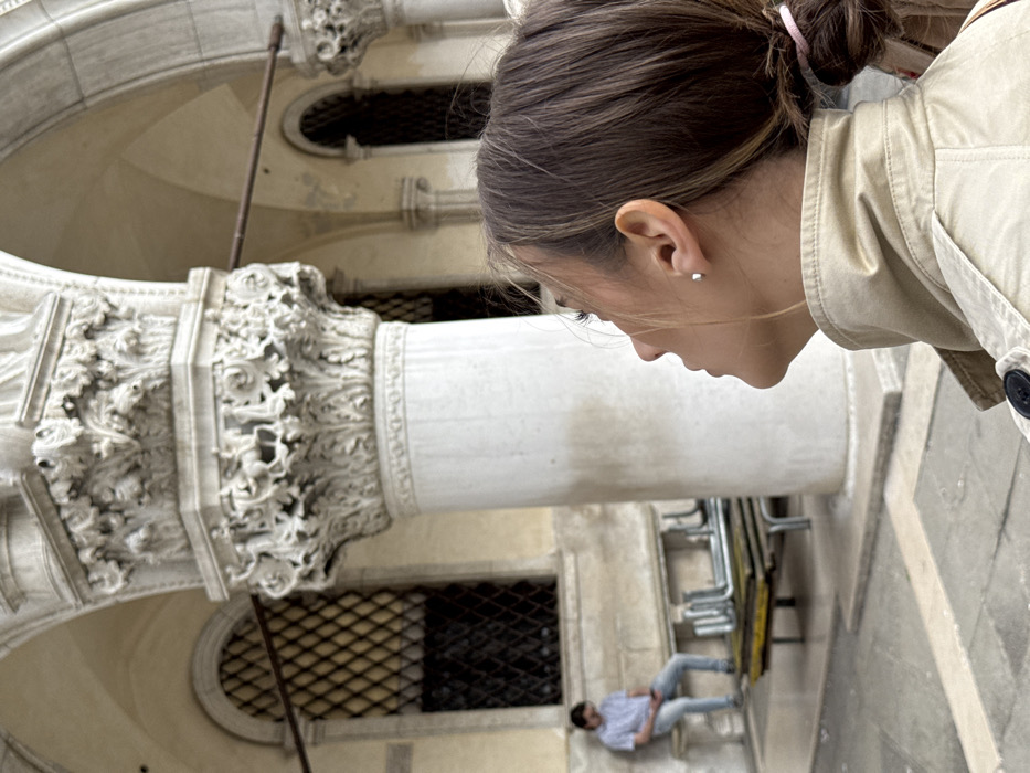 A three-quarter profile of a woman in a tan trench coat, with brown hair in a ponytail secured by a pink elastic, occupies the lower right foreground, looking upwards and to the left. She wears a silver stud earring in her visible ear. Behind her, a prominent white marble column with intricately carved capitals rises, featuring zoomorphic and foliate designs. In the midground, to the left of the column, a man in a light blue striped shirt and light trousers sits on a stone bench, looking down at a mobile phone. Stacked metal frames with yellow bases are visible on the ground in front of the bench. The background displays further architectural elements typical of the Doge's Palace in Venice, Italy, including additional ornate columns, pointed arches, and arched windows with dark metal grilles. The ground is paved with large stone slabs.