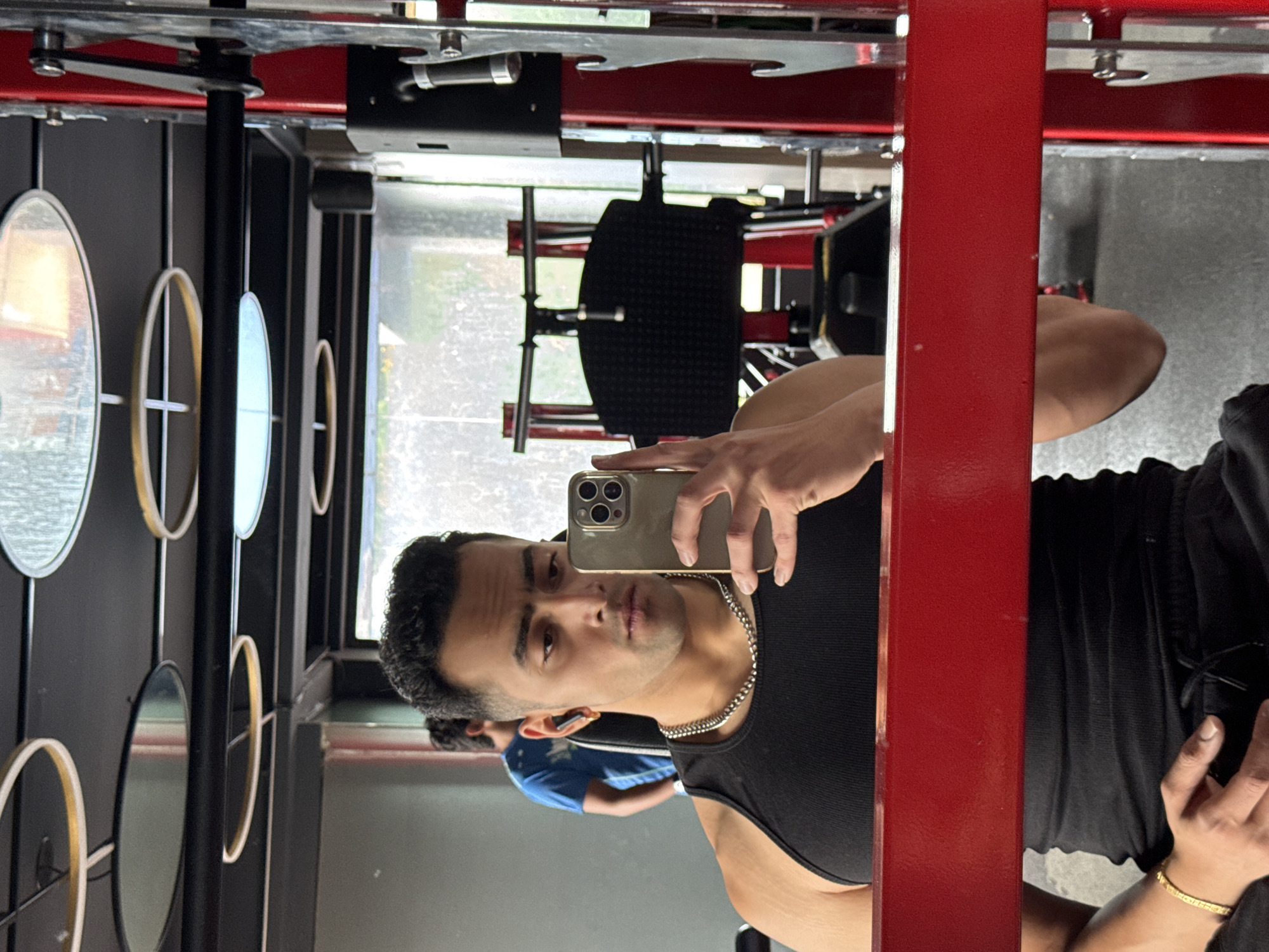 A man is taking a selfie in a gym mirror, the scene is rotated 180 degrees. The man is wearing a black tank top and a silver chain necklace. He's holding a silver smartphone. In the reflection, a man in a blue shirt is partially visible. Behind him is a weightlifting machine. The gym's décor includes a wall with circular decorative elements and a window. There is insufficient information to determine if the location is in Kolkata, India.