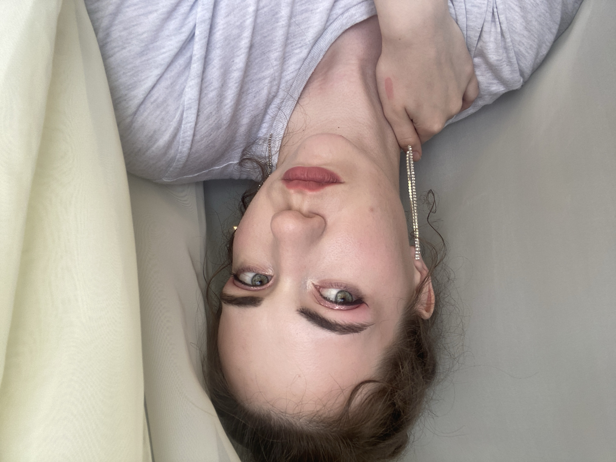 This is a close-up, overhead, and rotated photo of a young person lying down. They are wearing a light grey t-shirt and have makeup on, including pink lipstick and eyeshadow. A dangling earring with rhinestones is visible on their right ear. The person is lying on a light grey surface, with a sheer, pale yellow fabric draped to their left. The lighting is soft and diffused, suggesting it might be daytime indoors, as there are no strong shadows. The person's expression is neutral. The background is plain and undetailed, focusing attention on the individual. No text is visible in the image. The location context of Zavodoukovsk, Russia, does not provide any specific visual cues within the image itself.