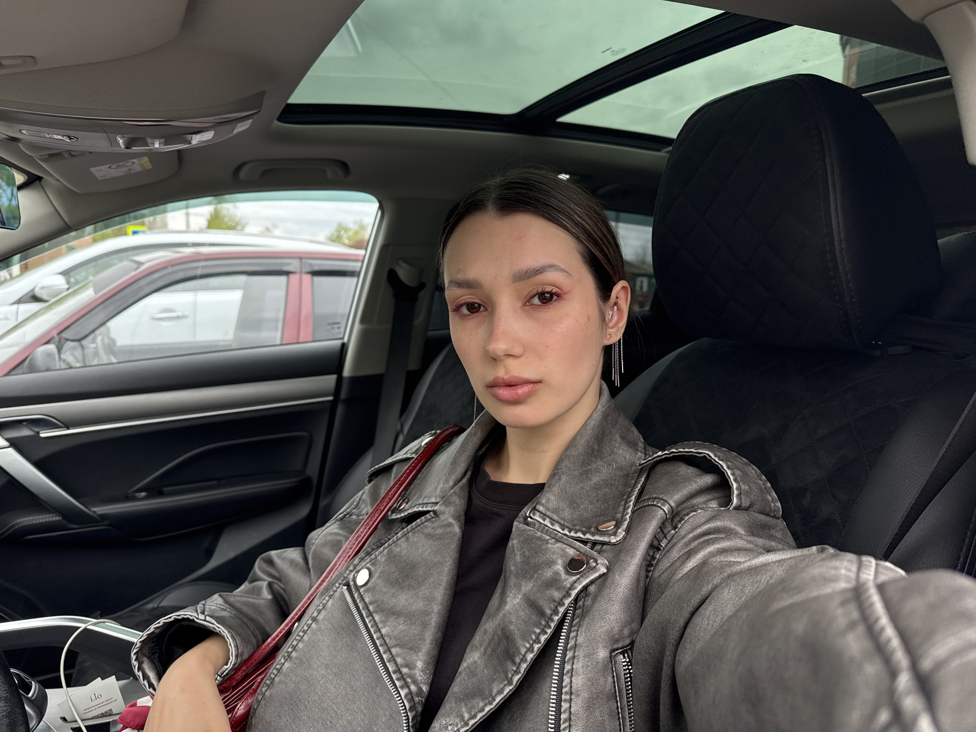 A woman is sitting inside a car, visible from the chest up. She has brown hair pulled back, fair skin, and is wearing a grey leather jacket over a black shirt, with a red bag over her shoulder. She has earrings in her left ear and is wearing red makeup around her eyes. The car has a sunroof that is open and it is cloudy outside. Other cars are visible through the car windows.