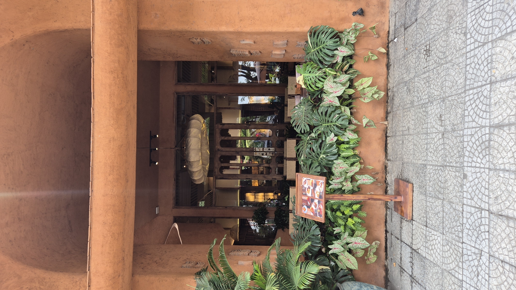 This is a medium-shot, eye-level outdoor photograph of the entrance to a restaurant in Da Nang, Vietnam. The building facade is painted a warm, earthy orange. The entrance features large, dark wooden double doors with glass panels, revealing the interior of the restaurant where tables and chairs are visible. Above the doors, a large, textured, white pendant lamp hangs. In front of the entrance, a wooden signpost displays a menu with images of food. A dense arrangement of lush green plants, including large monstera leaves and smaller plants with red-veined leaves, lines the base of the building and surrounds the signpost. To the left, a portion of a large potted plant with feathery fronds is visible. The foreground is a sidewalk paved with light grey square tiles arranged in a circular pattern. The overall impression is one of a welcoming and naturalistic dining establishment.