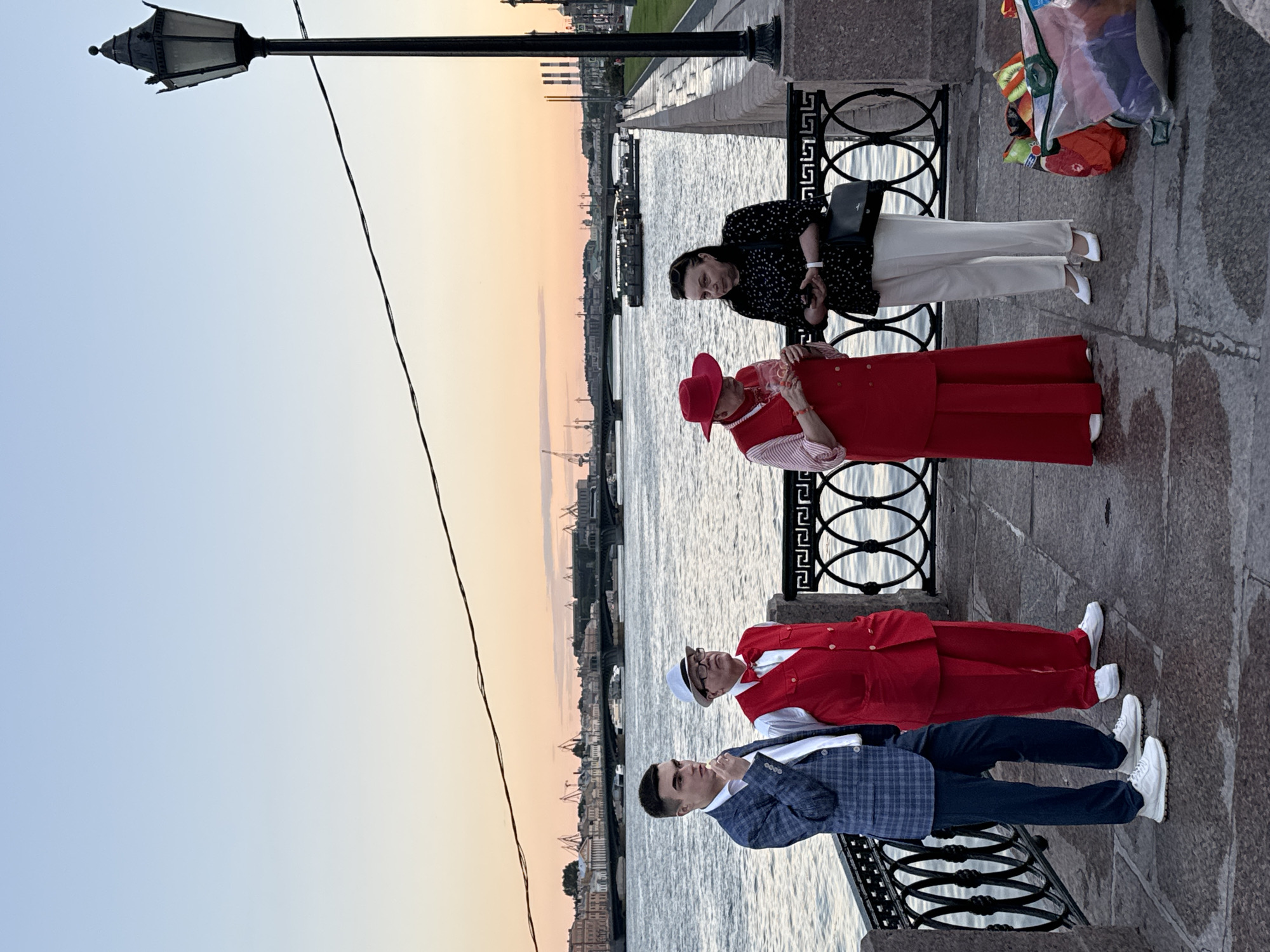 Four individuals stand on a stone embankment overlooking a wide river in Saint Petersburg, Russia, during sunset. The sky displays a gradient of soft blues and oranges. A vintage-style lamppost stands to the right, and a power line stretches across the upper portion of the frame. In the background, a cityscape with buildings and bridges is visible across the water.
From left to right, the individuals are:
1. A young man wearing a blue checkered blazer, a white shirt, and dark trousers, with white sneakers. He is looking down and to his right, with his left hand near his mouth, possibly holding something small.
2. A man in a red vest, red bow tie, white shirt, and white hat with glasses. He is looking forward.
3. A person wearing a wide-brimmed red hat, a red vest over a striped shirt, and long red trousers, with white shoes. They are holding a glass containing a pink liquid.
4. A woman in a black patterned top, cream-colored trousers, and white heels. She is holding a smartphone in her right hand and has a black purse on her left shoulder. She is looking at her phone.
A decorative black metal railing runs along the edge of the embankment in front of the group. To the right, near the woman, a collection of colorful bags and objects are piled on the ground. The pavement is made of stone slabs, and there are some puddles, suggesting recent rain. The overall atmosphere is calm, with the setting sun casting a warm glow over the scene.