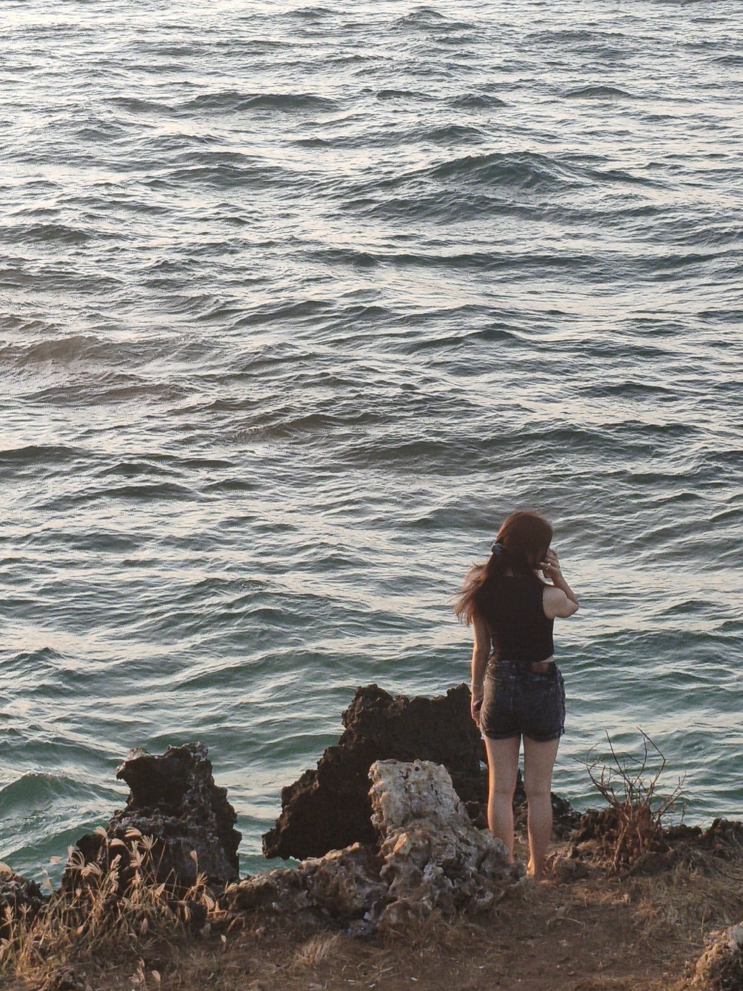 A young woman stands on a rocky shore, facing the ocean. She has long, brown hair, which is slightly windswept, and is wearing a dark-colored sleeveless top and denim shorts. Her back is to the viewer. The ocean is choppy, with small waves reflecting the light. The shore is covered in brown dry grass and rough, dark volcanic rocks. The scene appears to be a coastal area in Kuta Selatan, Indonesia, possibly during the late afternoon or early evening, as indicated by the lighting.