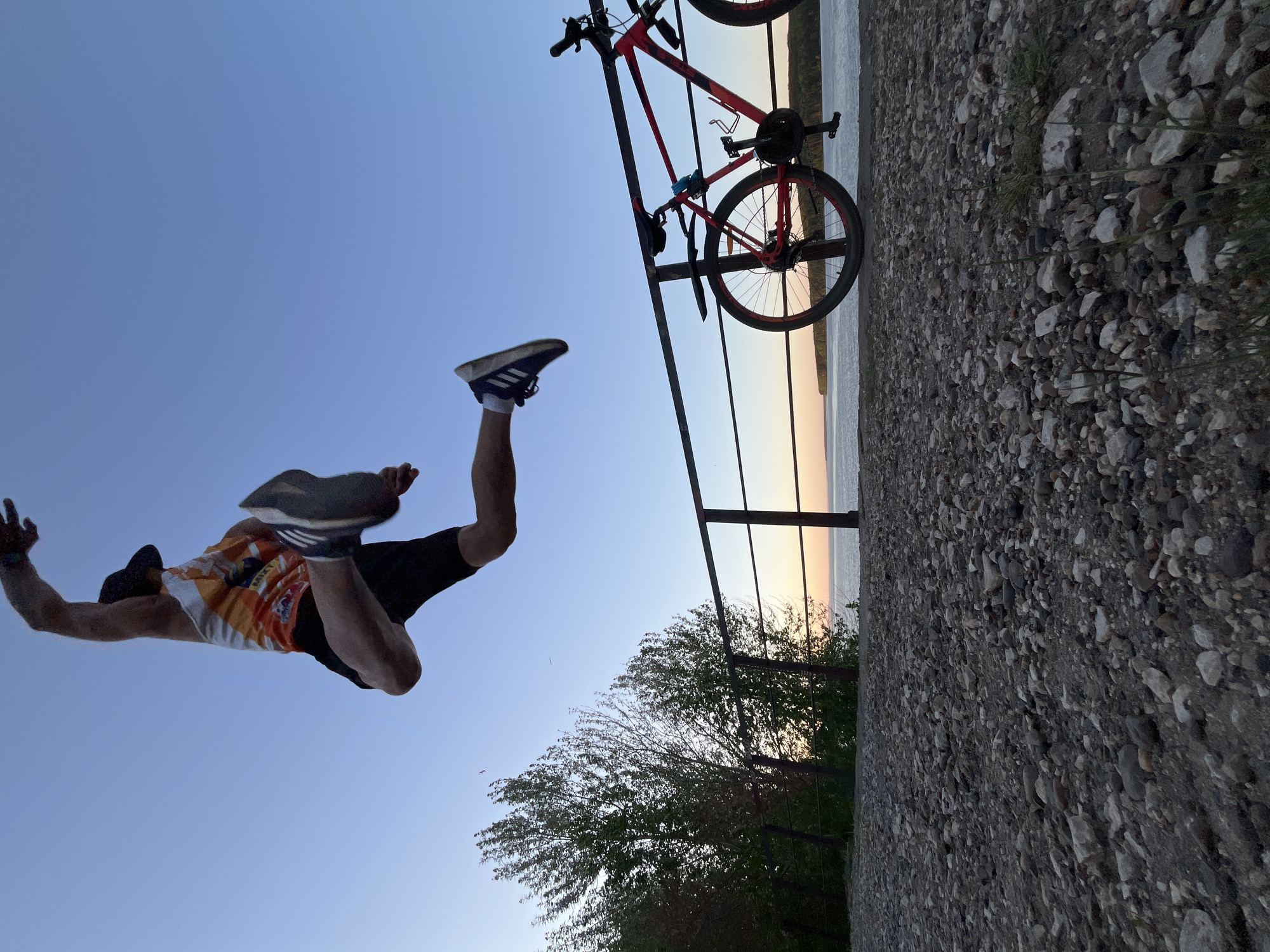 The image, taken in what is likely the city of Chaikovsky, Russia, shows a man in mid-air against a clear sky. He is wearing a cap, an orange and white patterned tank top, black shorts, and Adidas sneakers. His arms are extended. Nearby is a red bicycle propped against a metal railing. The railing overlooks a body of water, with trees visible below. The foreground is a concrete surface covered in small rocks. The sunset is occurring in the background with an orange hue. The image is oriented in a way where the horizon appears vertical.