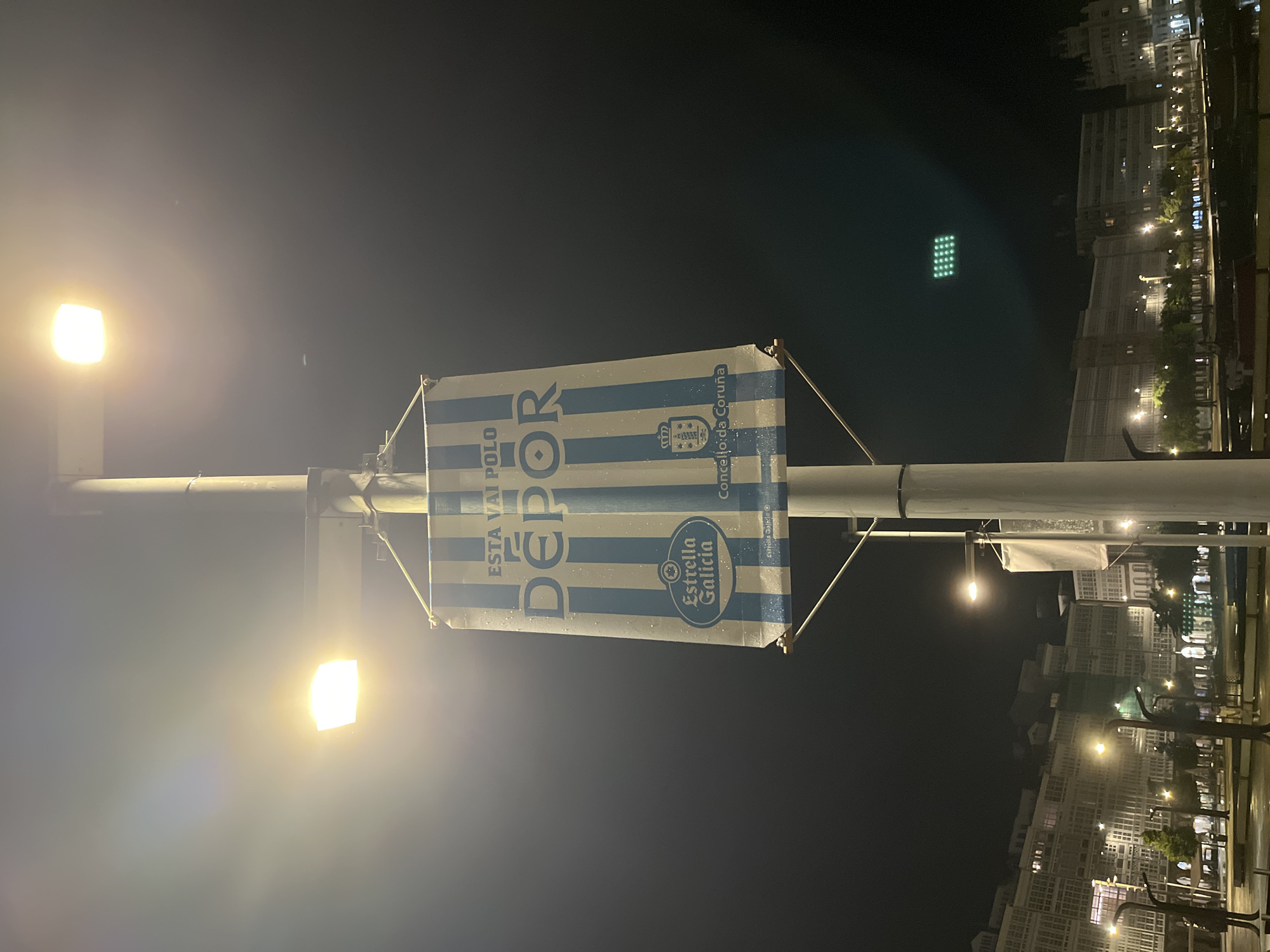 A street scene at night in A Coruña, Spain. A light pole with two illuminated lamps stands prominently. A banner attached to the pole displays blue and white stripes, along with text "DEPOR," "ESTA VAI POLO," and "Estrella Galicia," as well as the city's coat of arms. In the background, lit buildings and streetlights line the waterfront. A bright green grid of lights is visible further back, likely from a sports facility. The foreground is dark. The image is oriented vertically.