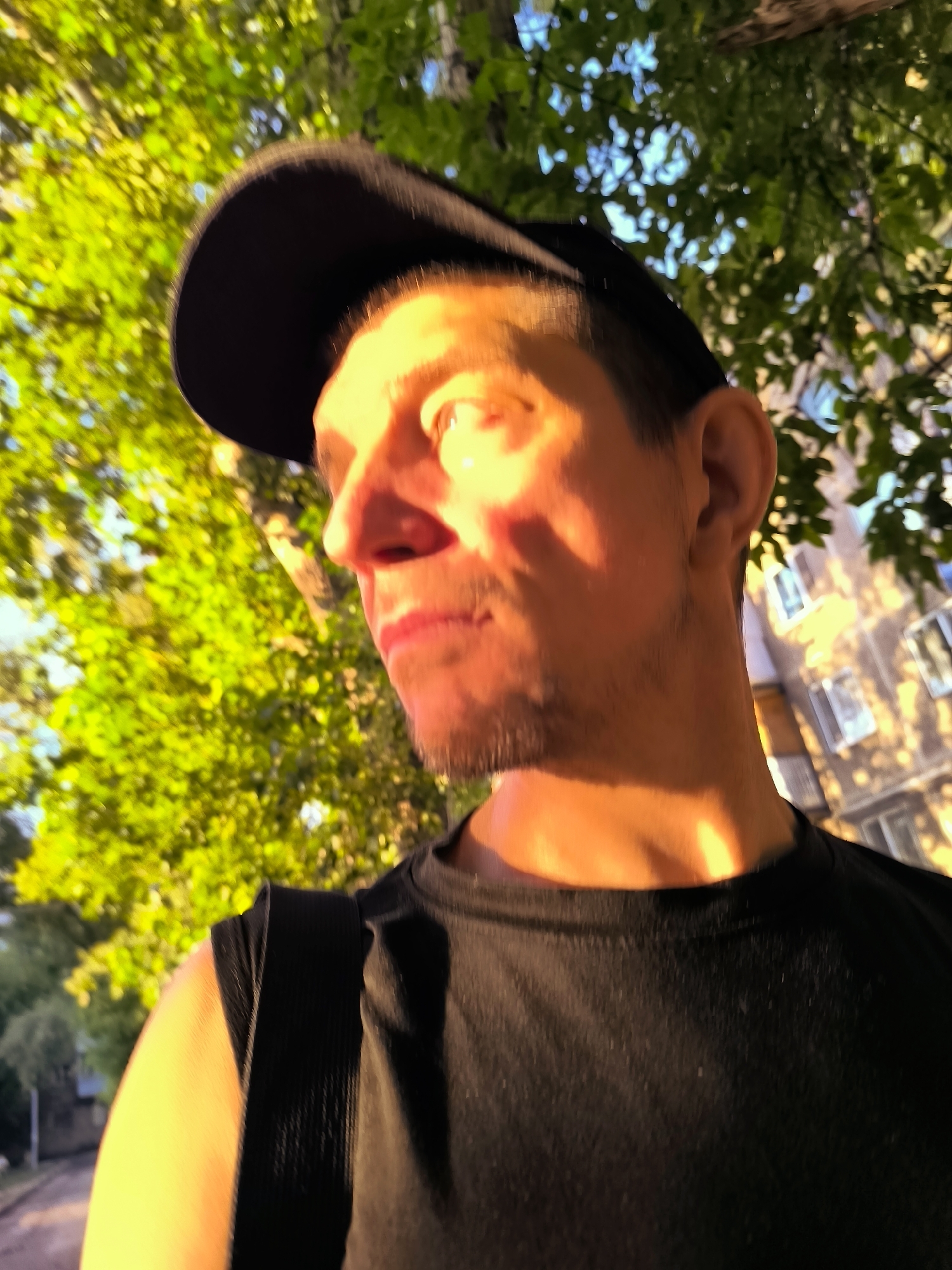 A man wearing a black t-shirt and a black baseball cap is outdoors, looking to the left. He has short, dark hair and stubble. The man is positioned in the foreground, with lush green trees and foliage behind him. In the background, a building with several windows is visible, illuminated by the warm light of the sun. The overall lighting suggests it is either early morning or late afternoon. The setting appears to be a residential area within Qarağandı city administration, Kazakhstan, with a visible paved pathway on the left side of the image.