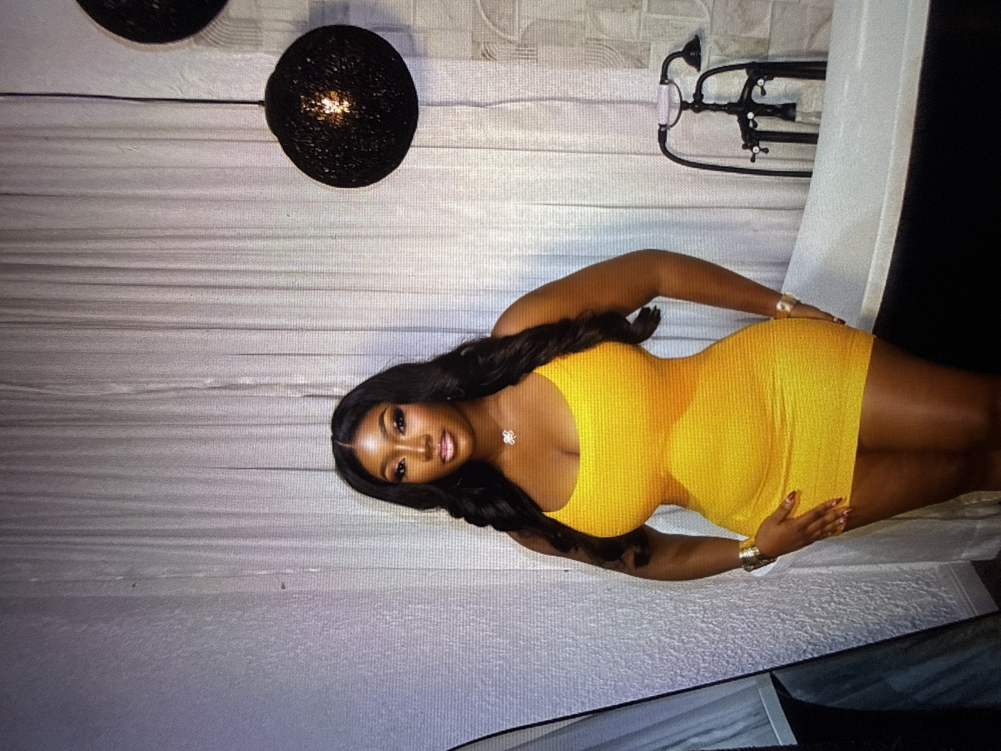 The media file shows a woman posing for a photo. She is wearing a bright yellow, form-fitting mini dress and has long, dark hair. She is also wearing a necklace with a flower-shaped pendant and a gold bracelet on her left arm. Her nails are painted red. The woman is positioned in what appears to be a bathroom or a similarly styled setting, indicated by the presence of a bathtub and what looks like a faucet or shower fixture on the wall behind her. The background features a textured, light-colored wall and a light-colored curtain or backdrop. There are also two black, spherical decorative items hanging from the ceiling in the upper left portion of the image. The lighting suggests an indoor setting, possibly during the day or evening. The woman's expression is confident and alluring. The overall mood is one of glamour and self-assurance. The context of Abuja, Nigeria, provides a geographical reference point for the image.