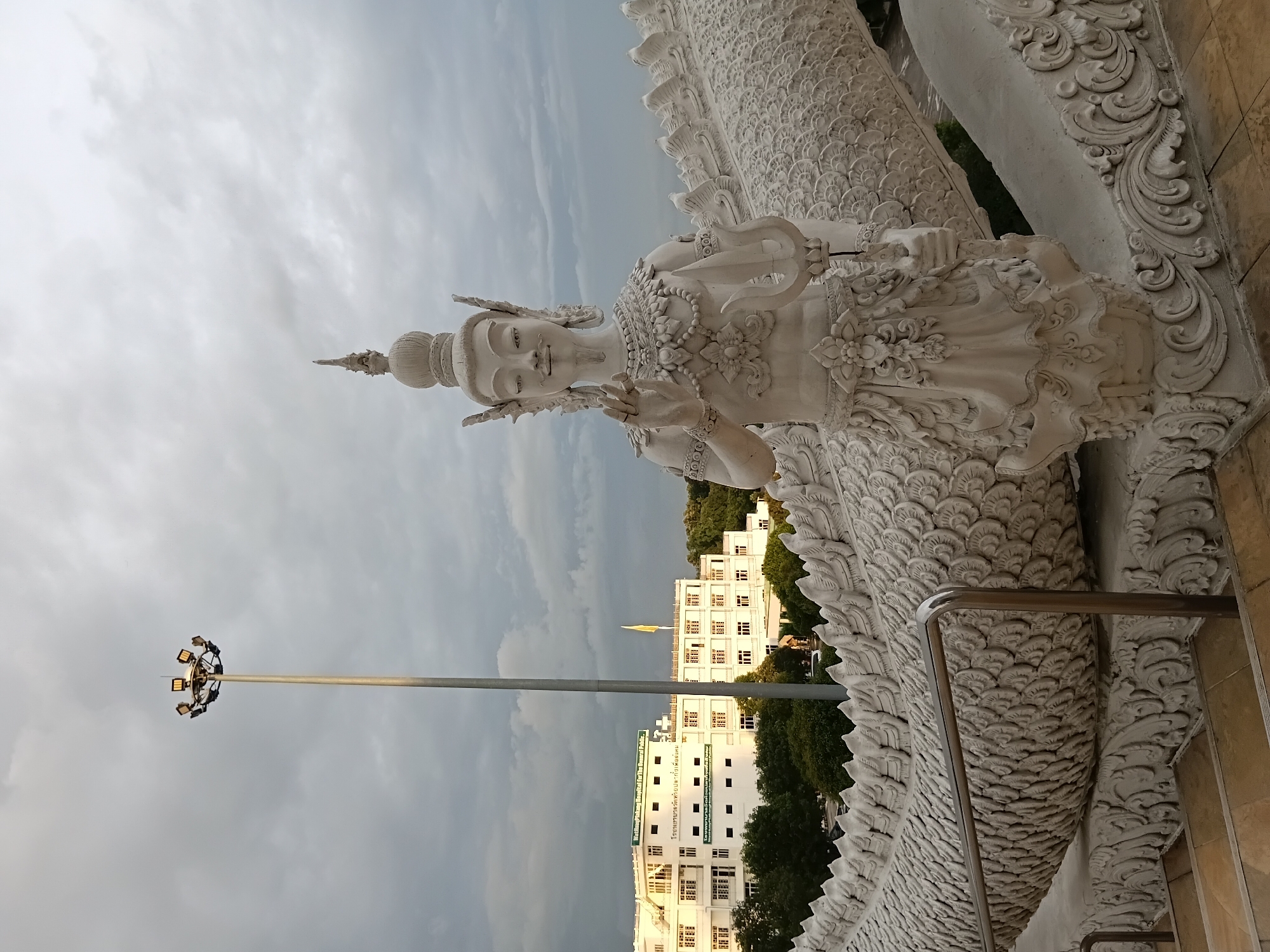 The image shows a white statue of a mythical being, possibly a deity or mythical creature, positioned in front of a large, textured serpent-like sculpture. The statue features ornate jewelry, a headdress, and an elaborate lower garment, with its right hand raised in a gesture of blessing or greeting. It holds a trident-like object in its left hand. The serpent sculpture has scales and decorative elements. In the background, a modern white building, identified by text as "Watthayong Hospital For The General Public," is visible, with trees and a tall lamppost. The sky is overcast with gray clouds. The foreground shows stairs and a metal railing. The scene appears to be outdoors, in an area of significant architectural and artistic interest.