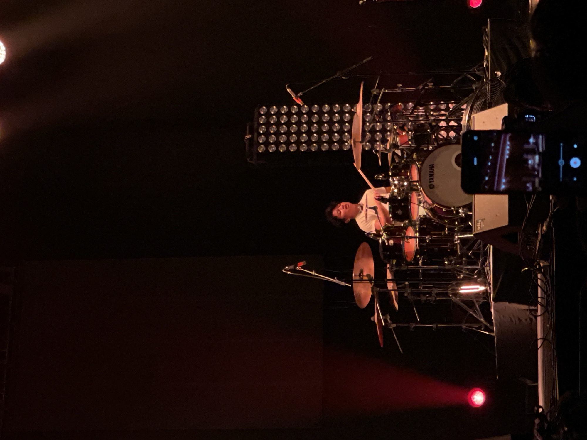 A drummer is seen performing on stage at a concert in Salembaran, Indonesia. He is playing a large Yamaha drum kit surrounded by cymbals. A stage light array is behind him. A person is holding a smartphone and recording the performance. A stage light is visible near the base of the stage.