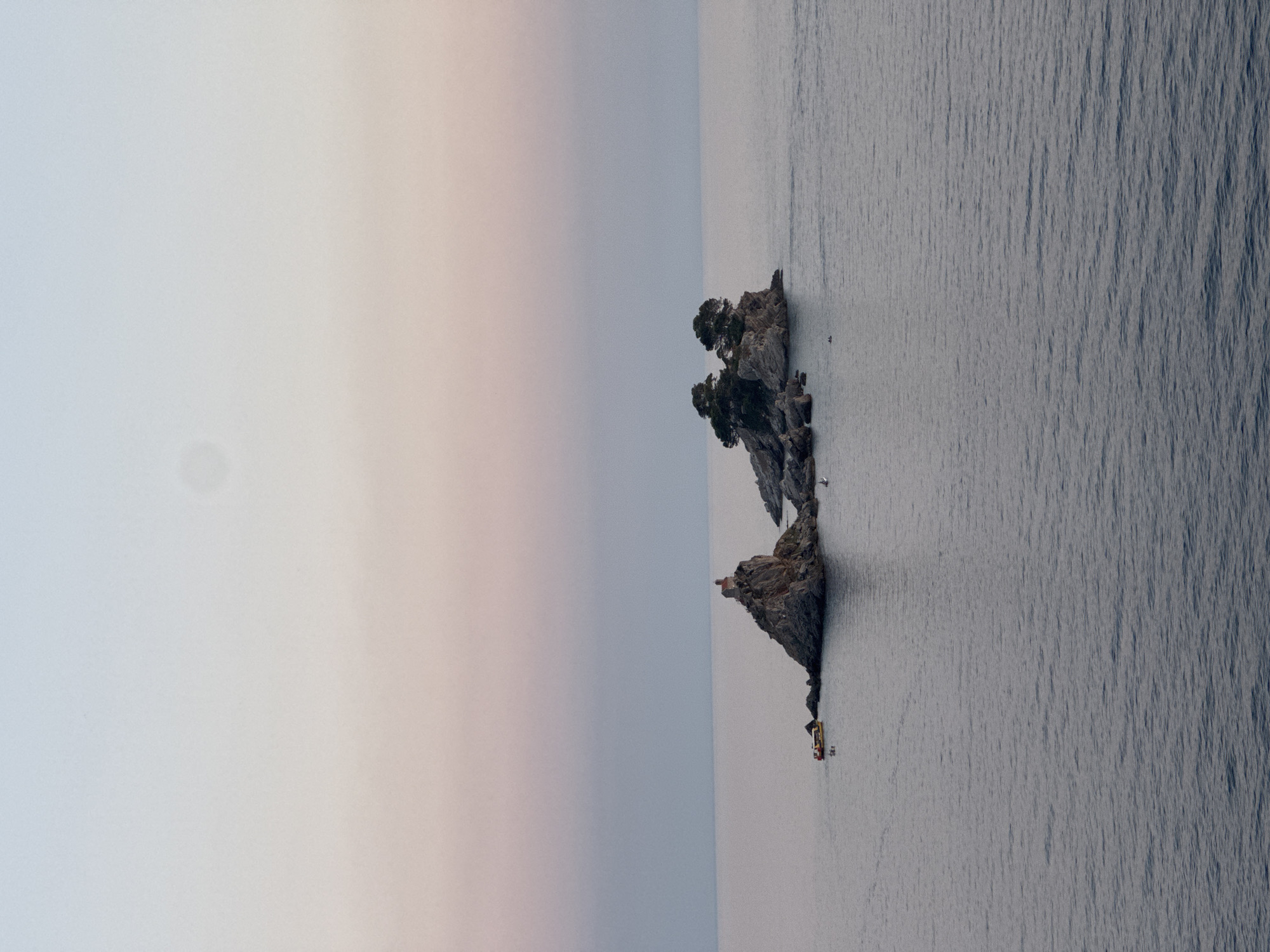 An eye-level, wide shot captures two rocky islets in the Adriatic Sea near Donji Katun, Montenegro. The islet on the left is topped with a small, red-roofed building, possibly a chapel or lighthouse. A yellow boat with people on board is anchored near this islet. The second islet, to the right, is covered in trees and smaller rocks. The sea is calm and reflects the soft hues of the sky, which transitions from a pale lavender at the horizon to a light blue at the top of the frame. A faint, circular shadow is visible in the upper portion of the sky.