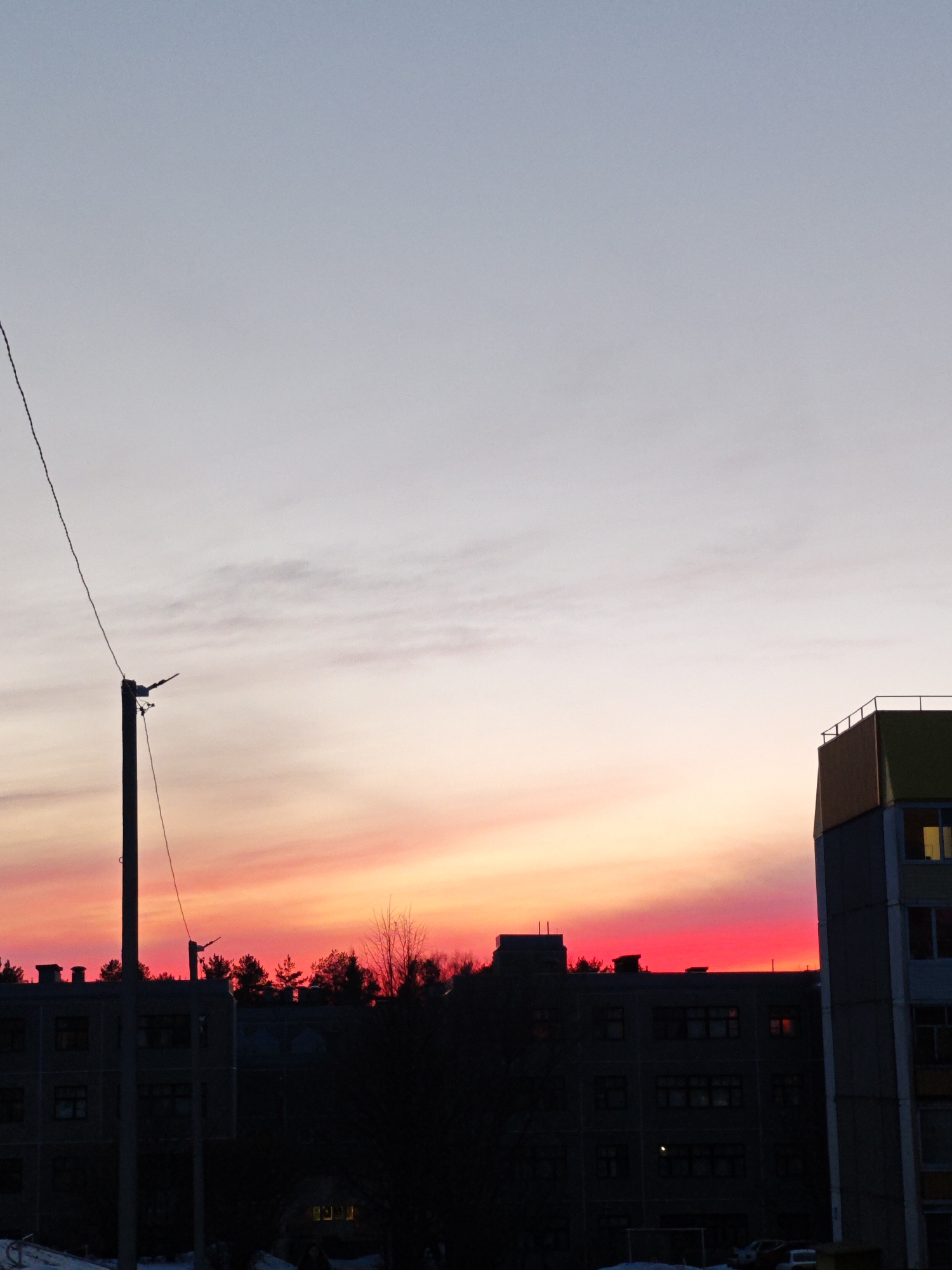 The image shows a sunset in what appears to be an urban environment, Марковский, Russia. A telephone pole stands on the left side of the frame, with wires extending toward the top. In the foreground, there are buildings silhouetted against the colorful sky. The sky transitions from light blue at the top to orange and red near the horizon. There are trees and more buildings visible in the midground, their shapes outlined by the vivid sunset colors. The buildings are dark and appear to be multi-story structures. Snow patches are visible in front of the buildings.