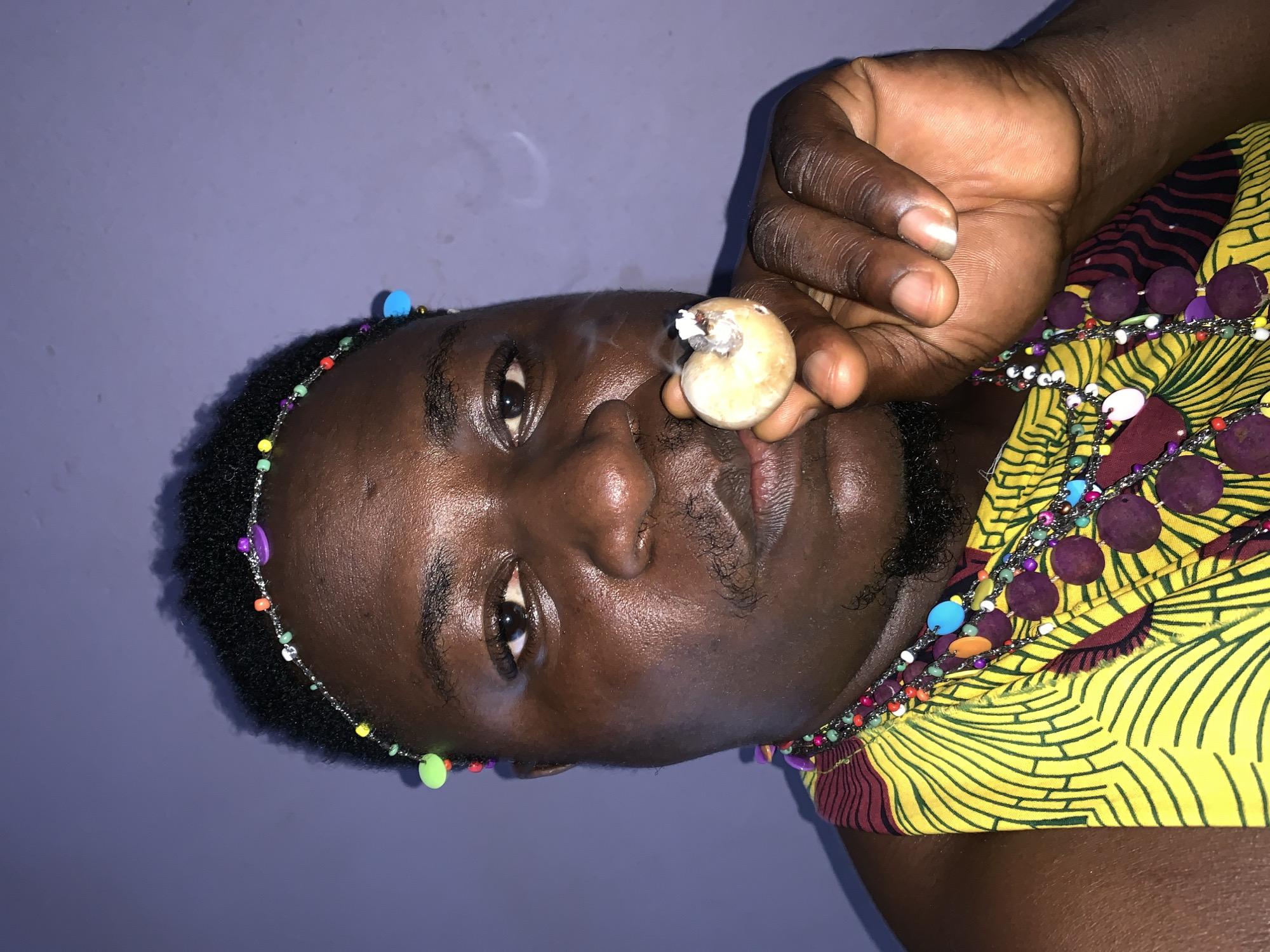 A close-up, medium-shot photograph captures a dark-skinned individual from the chest up, against a plain, muted purple background. The subject's head is turned slightly to their right, with their gaze directed forward. They are adorned with a colorful beaded headband and multiple beaded necklaces, including a prominent one featuring large, round, purple beads. They wear a vibrant yellow garment with a black and maroon abstract pattern. The individual is holding a lit object, possibly a cigarette or blunt, in their right hand, close to their face, with wisps of smoke emanating from it. The lighting in the photograph appears to be artificial, casting shadows and highlighting the texture of the subject's skin and the beaded accessories. The scene is indoors, given the solid colored backdrop and lack of natural light features. The setting cannot be definitively identified as Akwanga, Nigeria, based solely on the visual information provided.