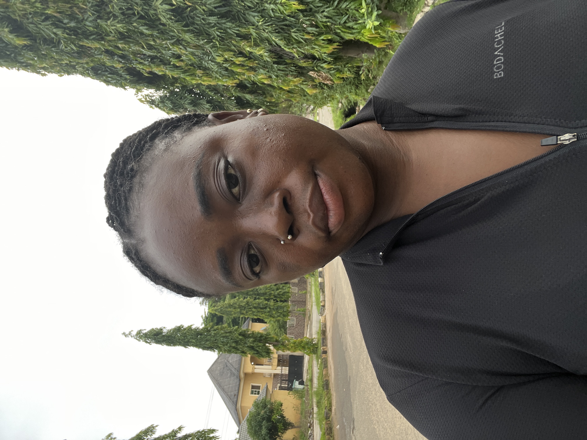 A medium-shot selfie captures a Black woman with braided hair, her face turned slightly towards the camera. She is wearing a black zip-up jacket with the logo "BODACHEL" visible on the right side. She has a nose ring and small earrings. The background shows a paved street, a yellow house with a dark roof, and several tall, slender trees. The overall environment suggests an outdoor setting in a residential area, likely in Abuja, Nigeria, given the architectural style and vegetation. The sky is overcast.