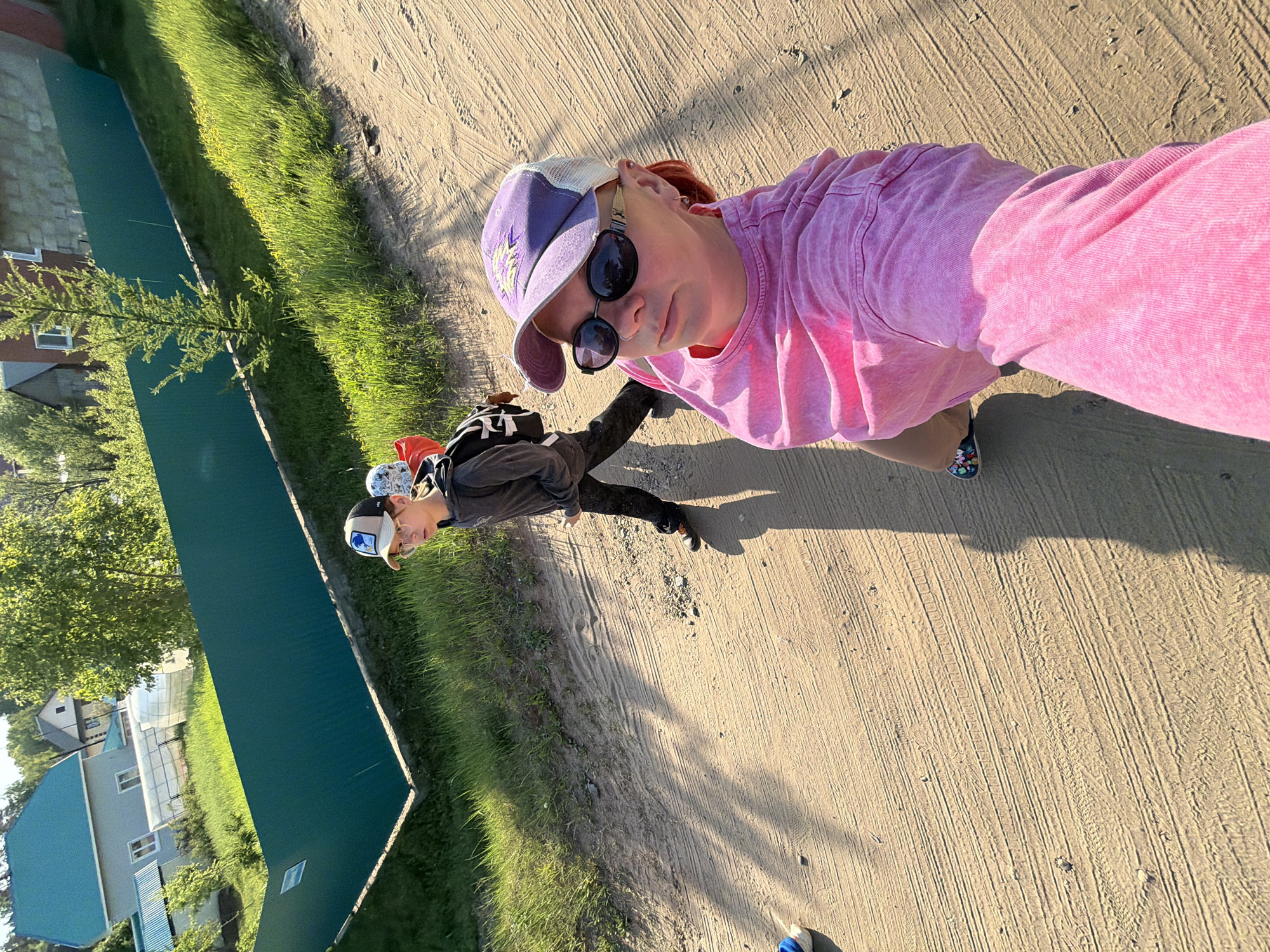 A selfie captures two individuals on a dirt road in what appears to be a suburban area in Chaikovsky, Russia. The person taking the picture is wearing a pink shirt and a purple baseball cap with sunglasses hanging from it. The second individual, slightly behind, is wearing a gray top, dark pants, a baseball cap, glasses, and a backpack.
The environment includes a green fence and lush vegetation on one side of the road, while houses are visible in the background. The dirt road itself is marked with tire tracks. The angle of the sun suggests it is either early morning or late afternoon.